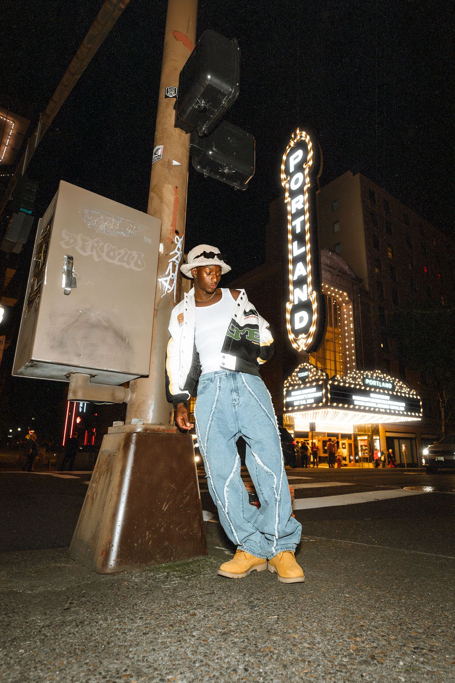 Young man in bucket hat, wide-leg jeans, and white jacket poses by a street pole near a lit theater marquee at night.