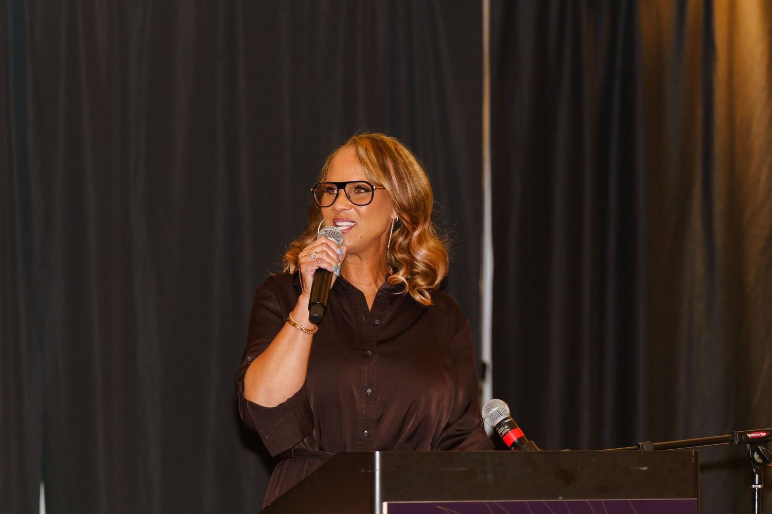 Woman with glasses speaking into microphone at podium during indoor event presentation.