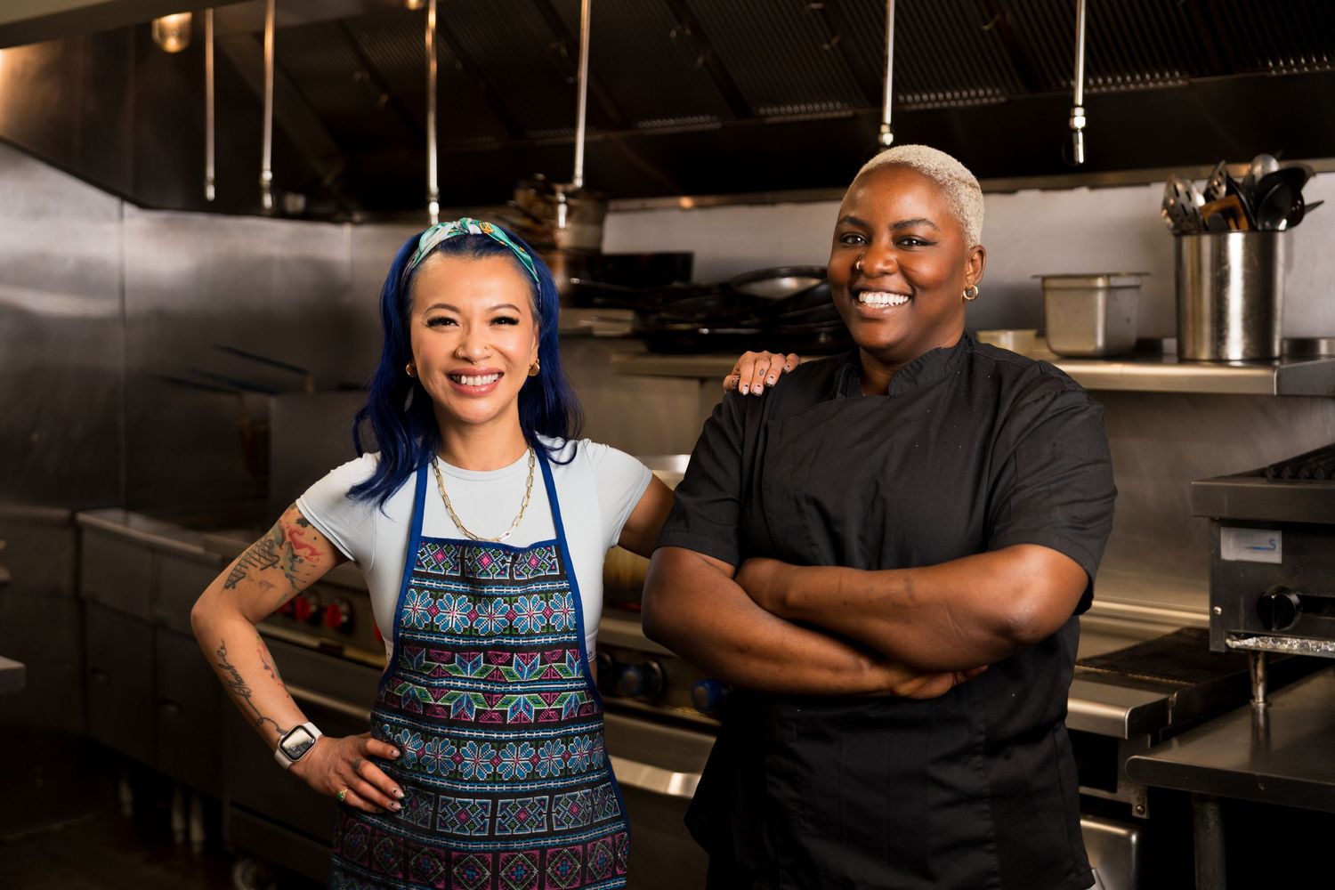 Two smiling chefs in aprons pose together in a professional stainless steel kitchen.