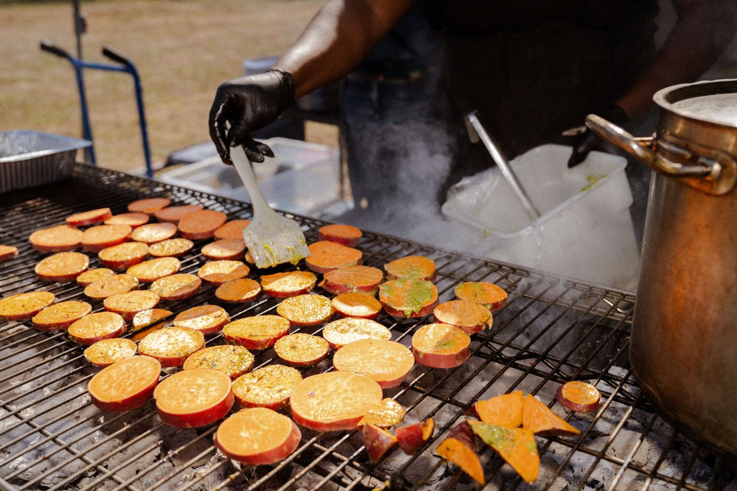 Sliced sweet potatoes grilling on an outdoor BBQ grill with steam rising, tended by a gloved chef.