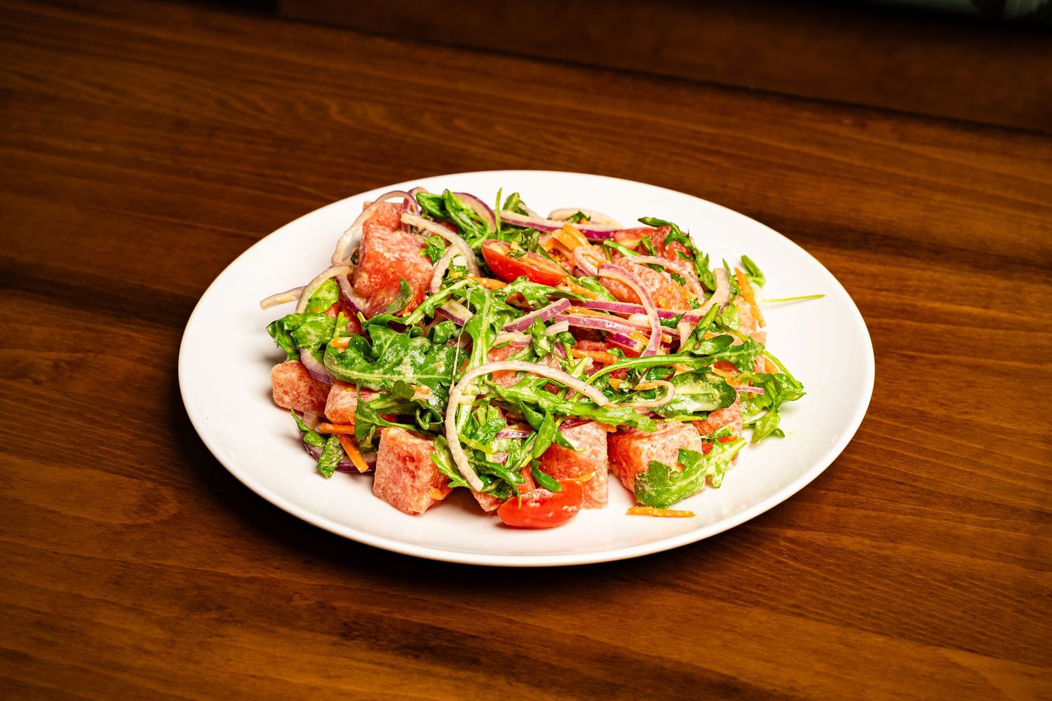 Fresh salad with arugula, tomatoes, onions, and meat on a white plate on a wooden table.