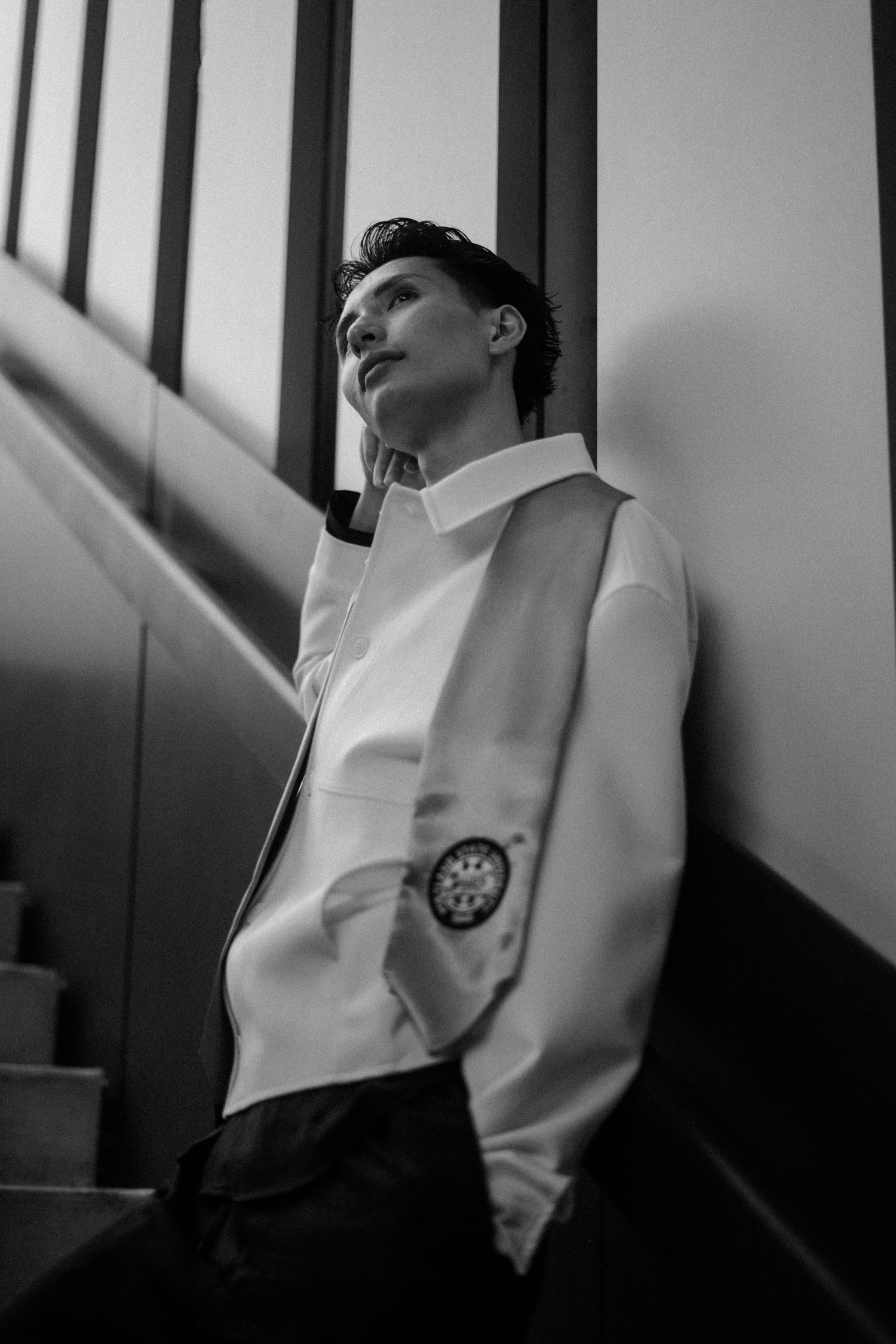Black and white portrait of a young man in a white shirt leaning against stairs, looking away thoughtfully.