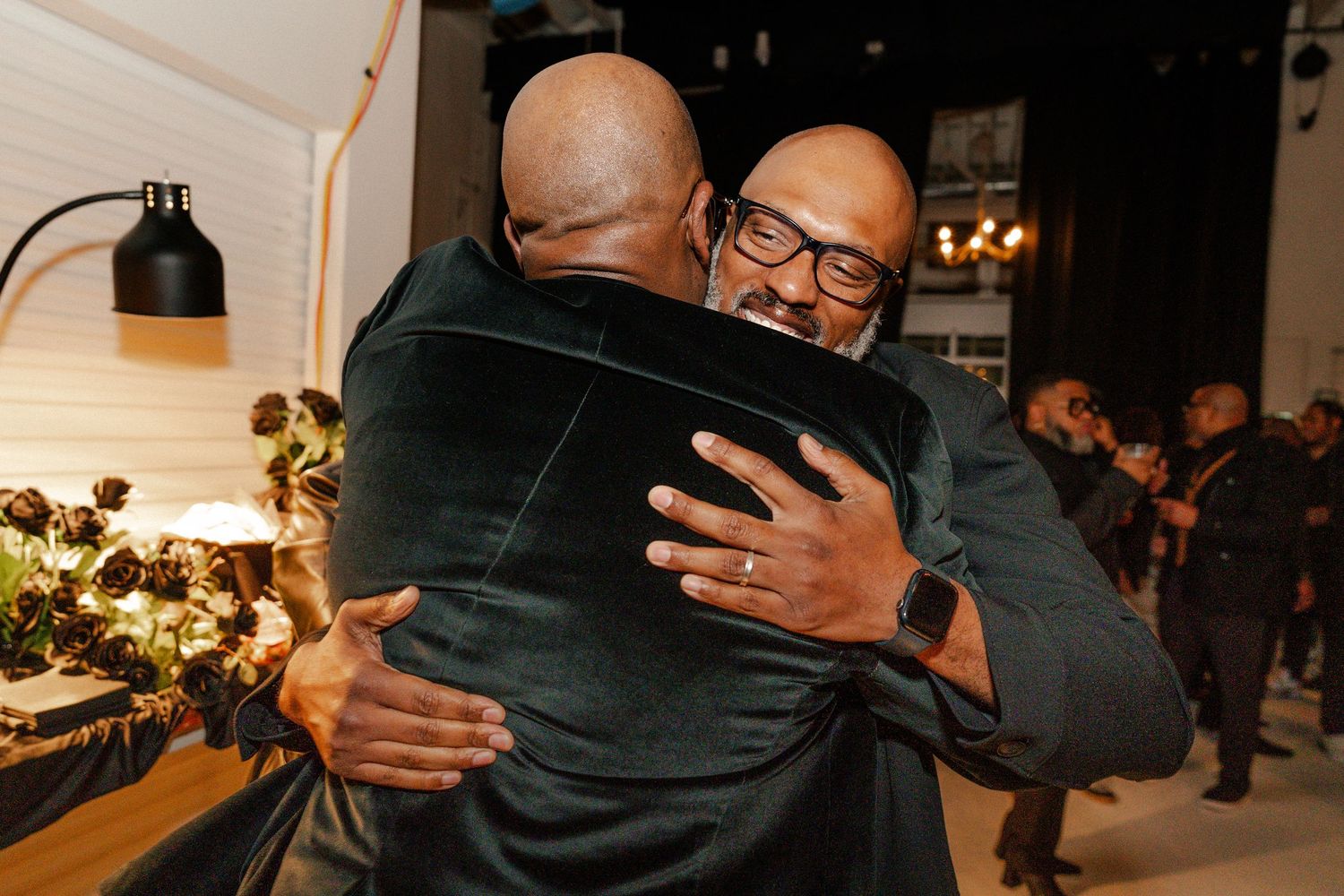 Two men in green velvet blazers share a warm hug at an elegant indoor event with floral arrangements.