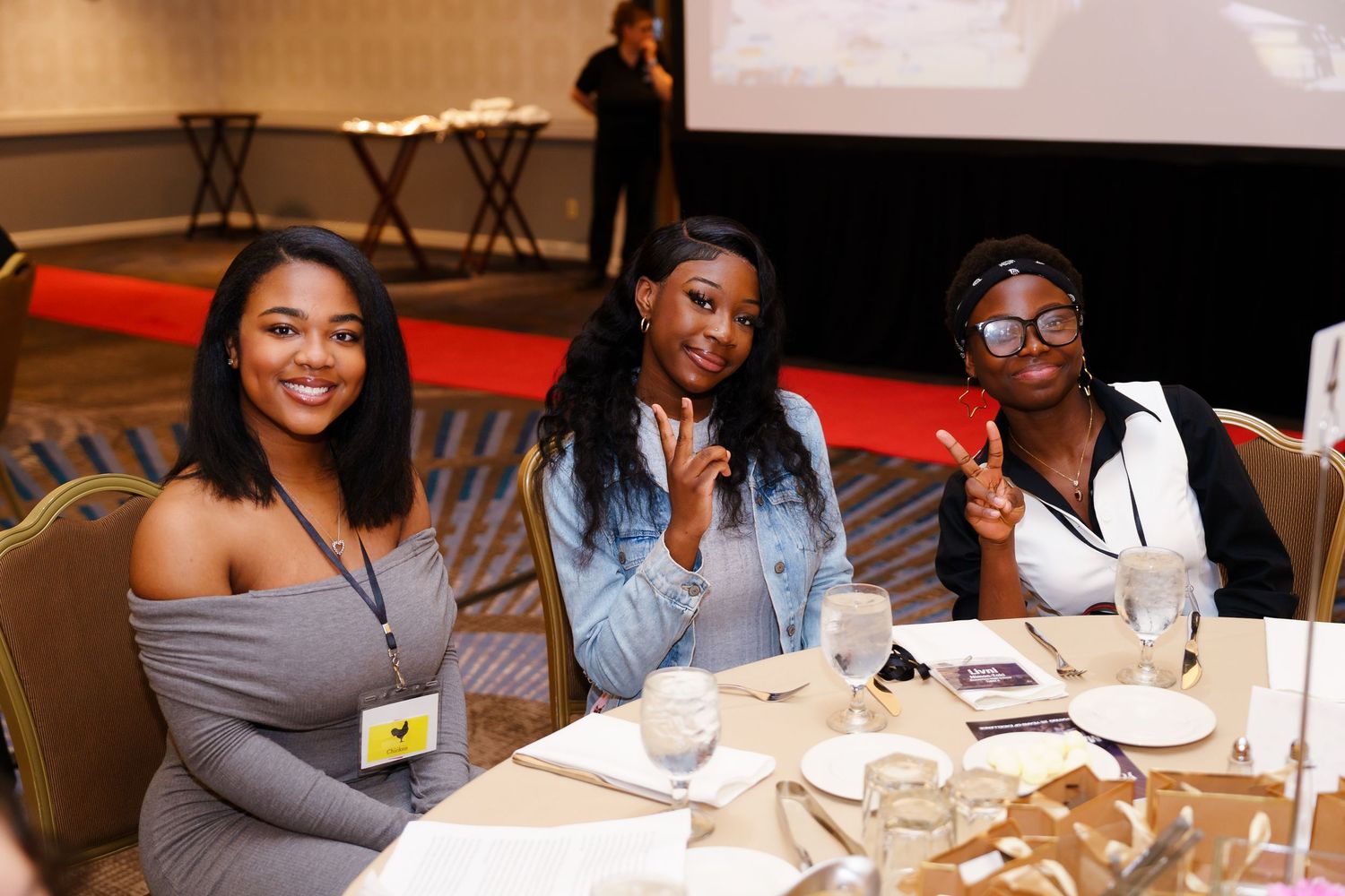Three young women smiling at a banquet table during a conference event in a hotel ballroom.