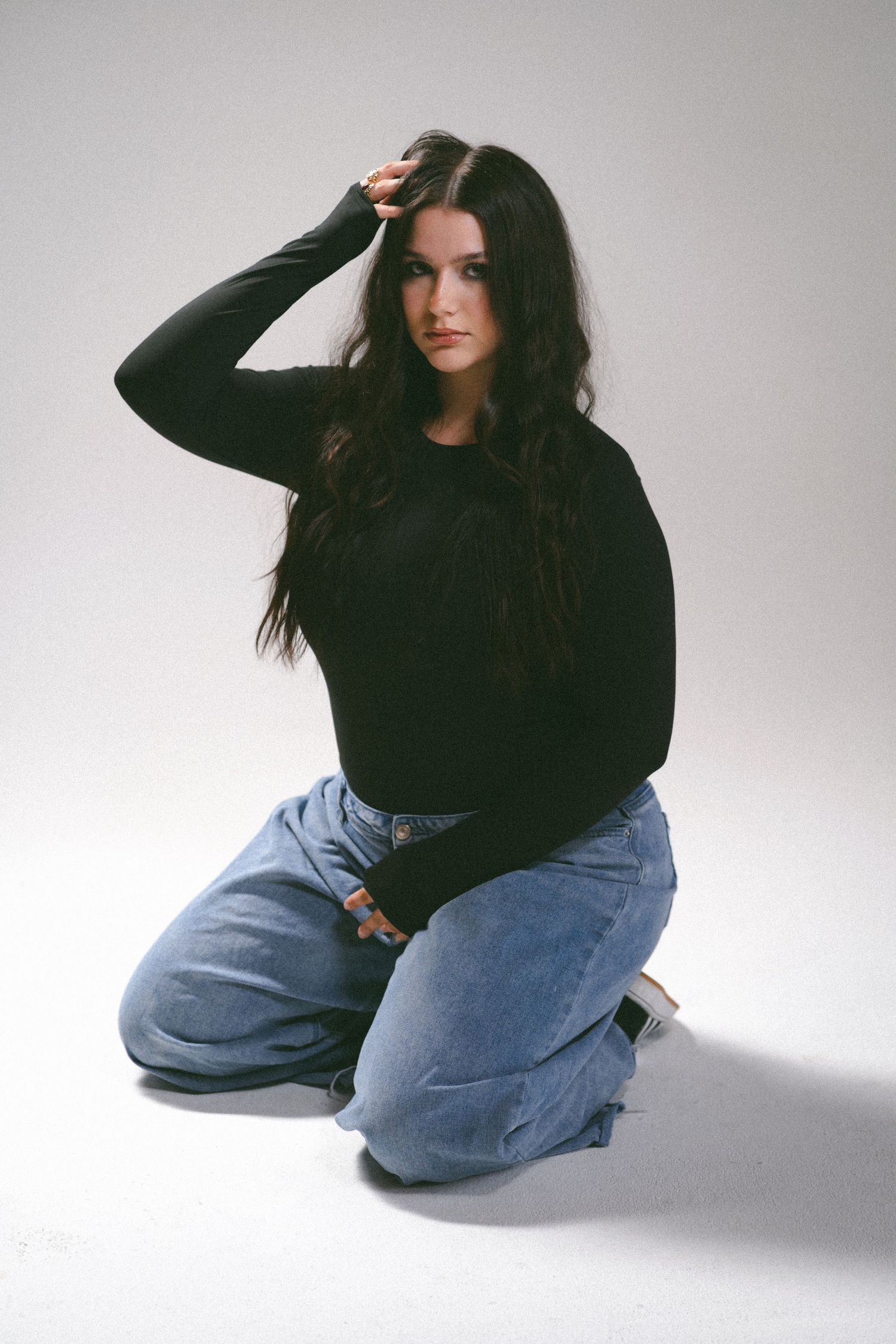 Young woman with long dark hair sits on floor wearing black sweater and blue jeans, posing for studio photo.