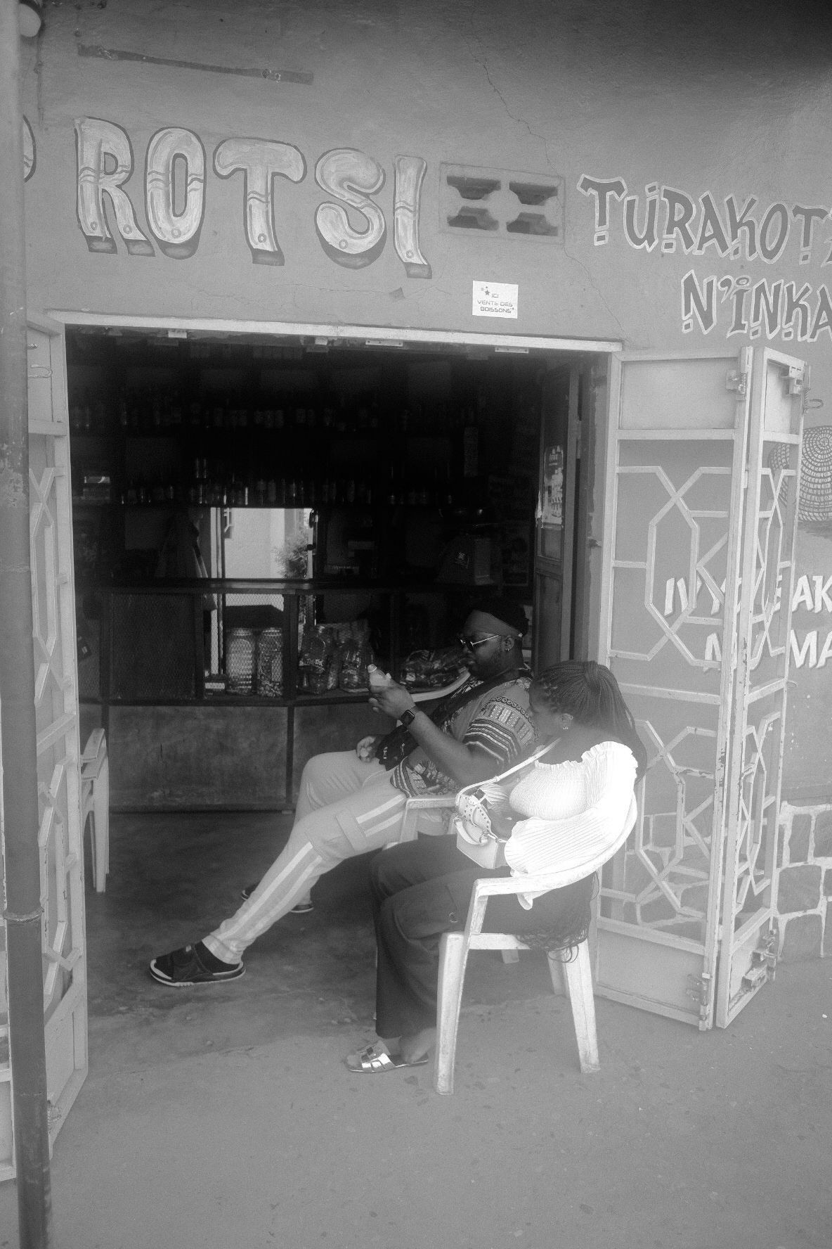 Black and white image of people relaxing in chairs outside storefronts with graffiti.