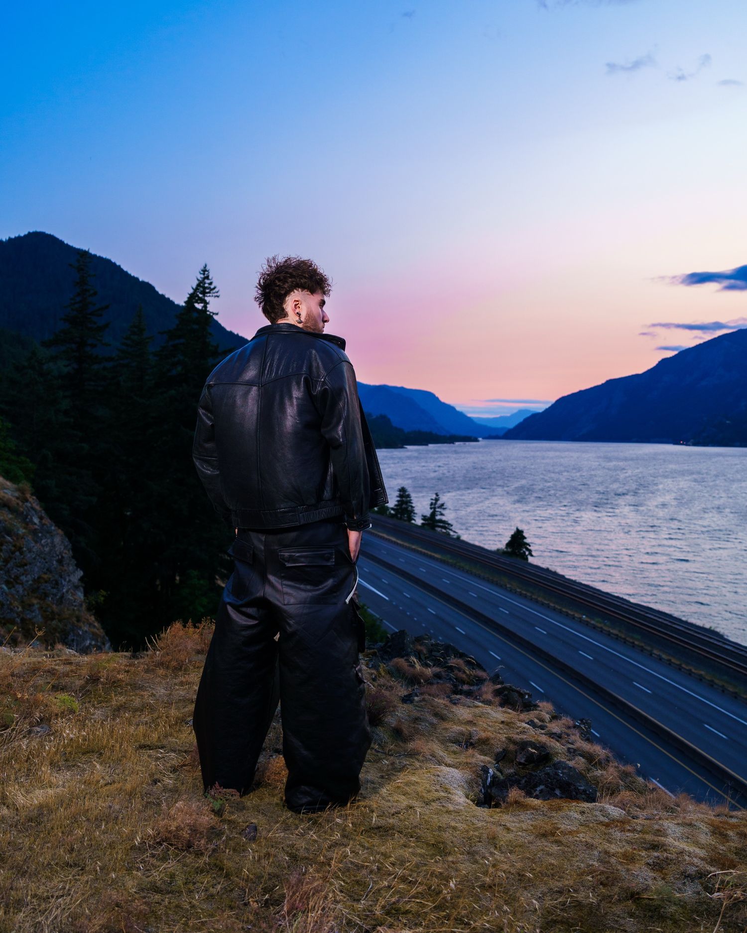 Person in black clothing sitting on rocky overlook at sunset near mountain lake.