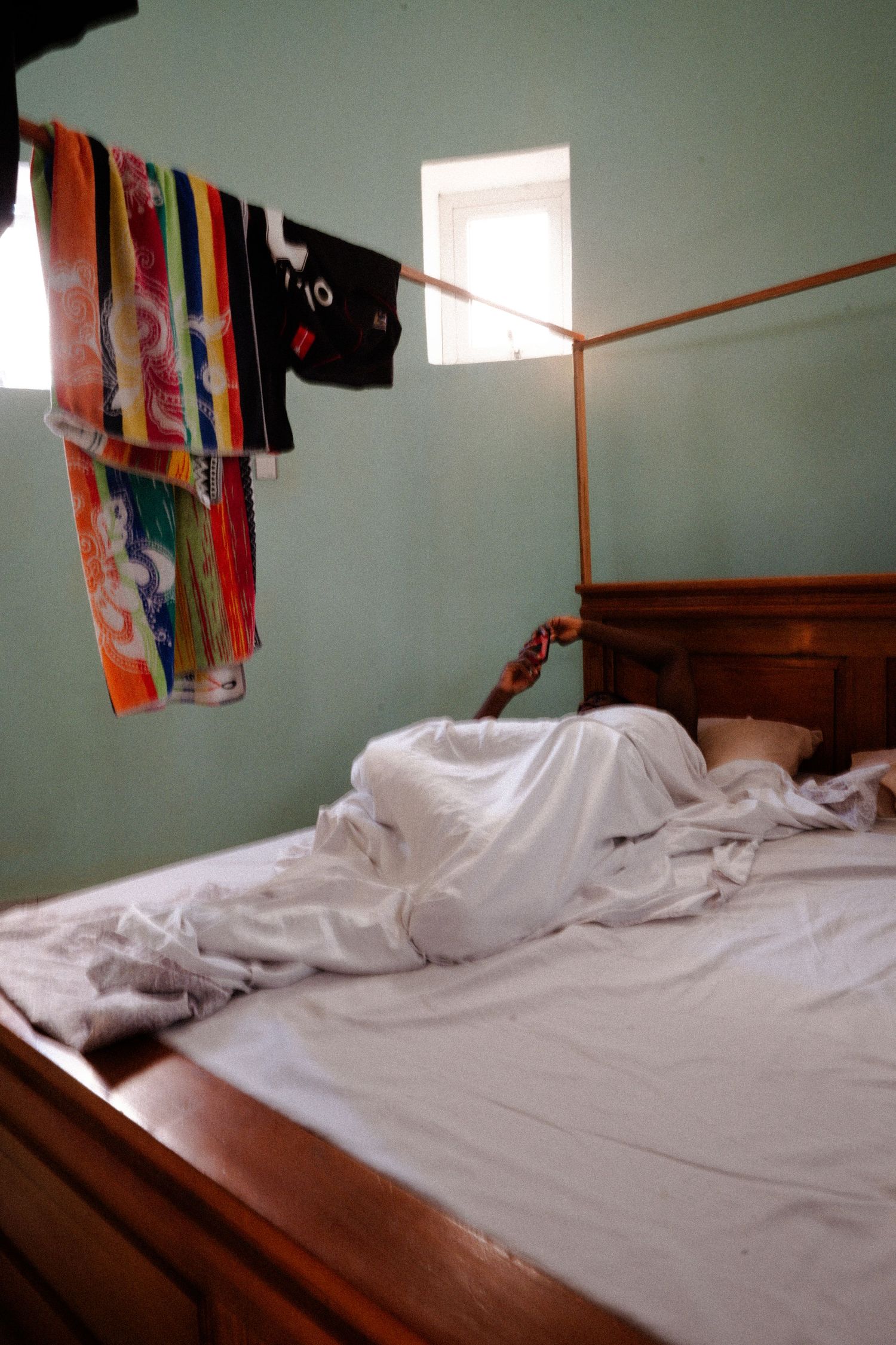 White bedding on wooden bed frame with colorful clothing hanging on line above.