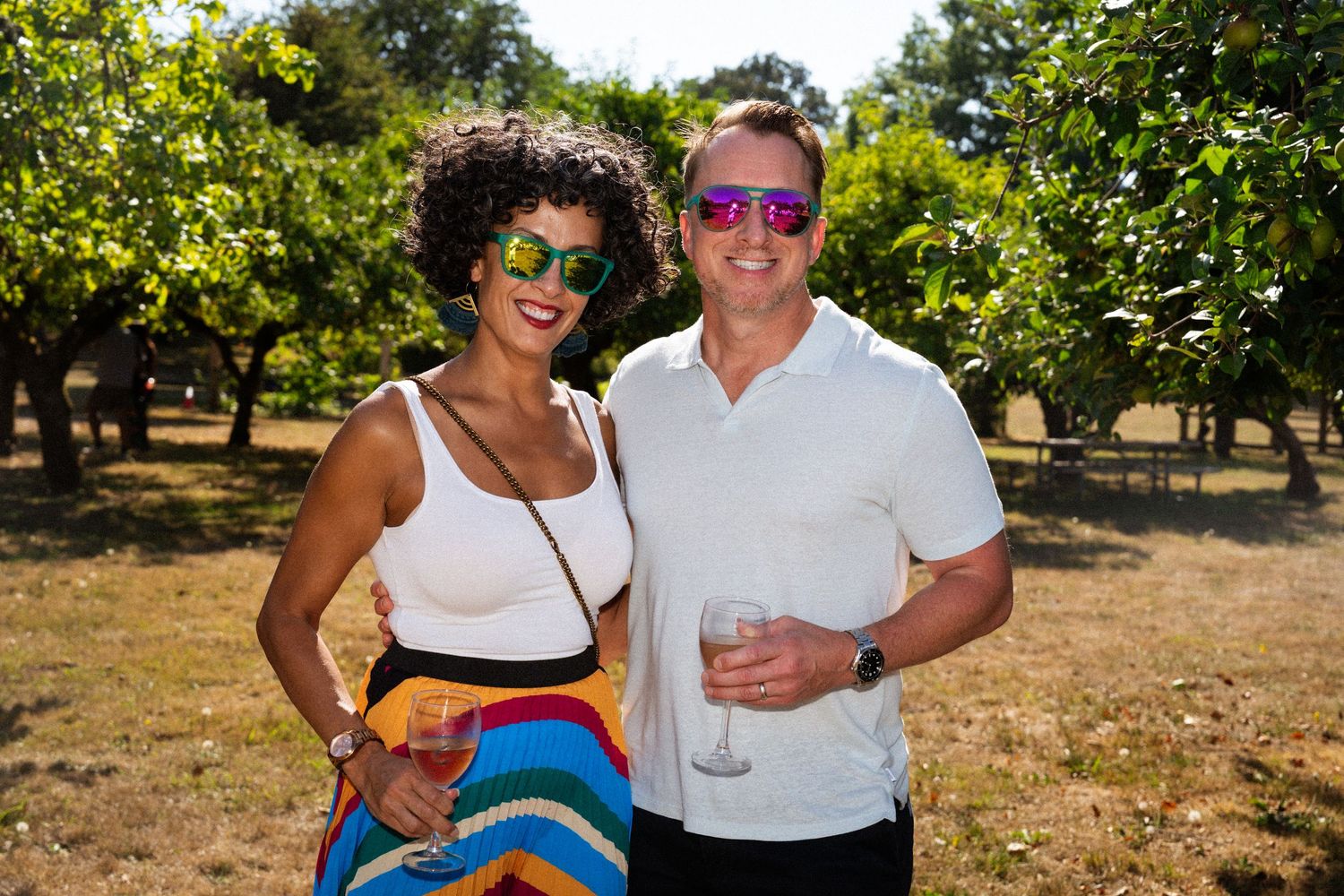 Smiling couple wearing colorful sunglasses enjoying wine in a sunny orchard vineyard setting.