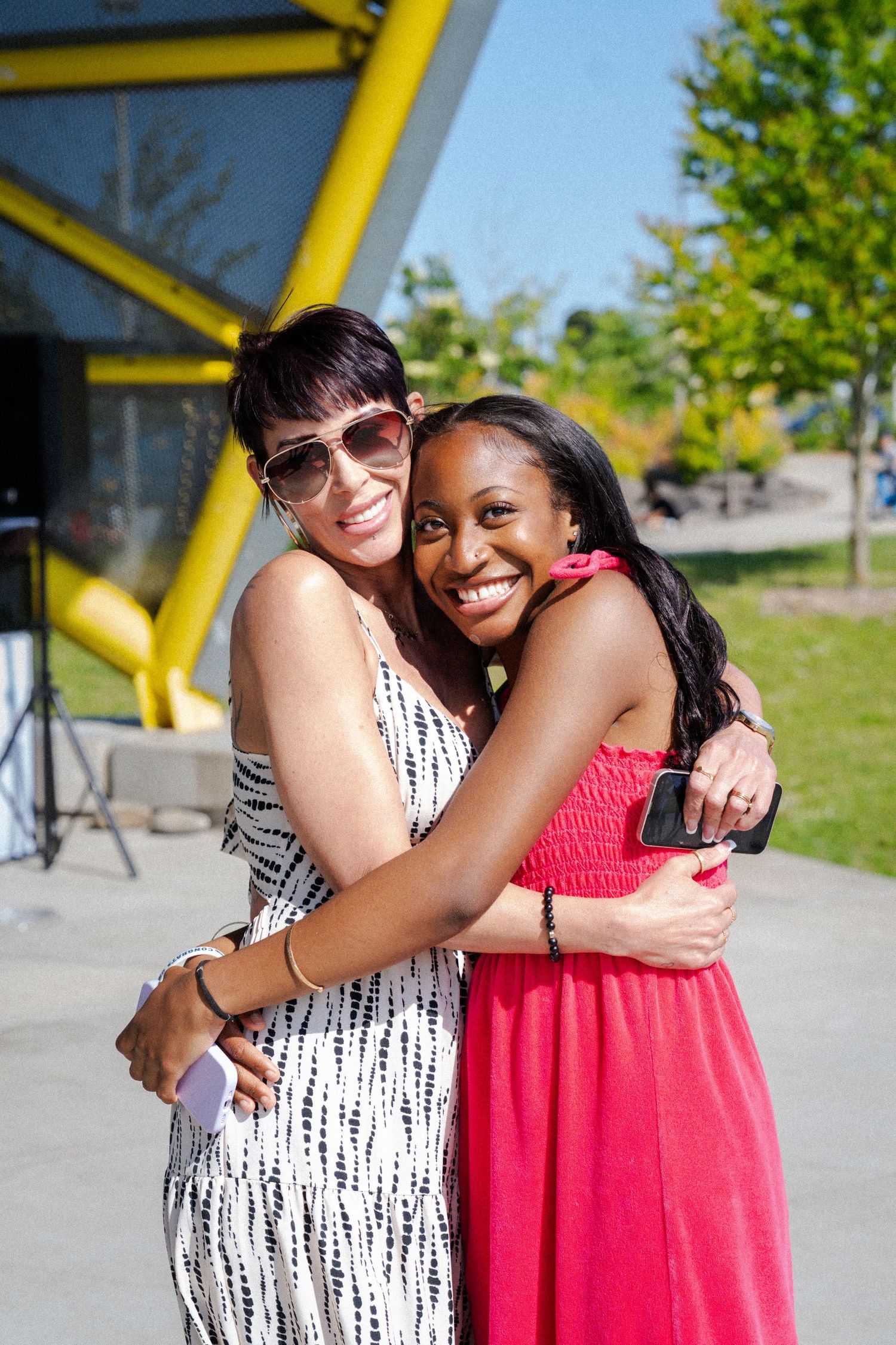 Two women smiling and hugging outdoors on a sunny day, one in a white dress and sunglasses, the other in a pink dress.