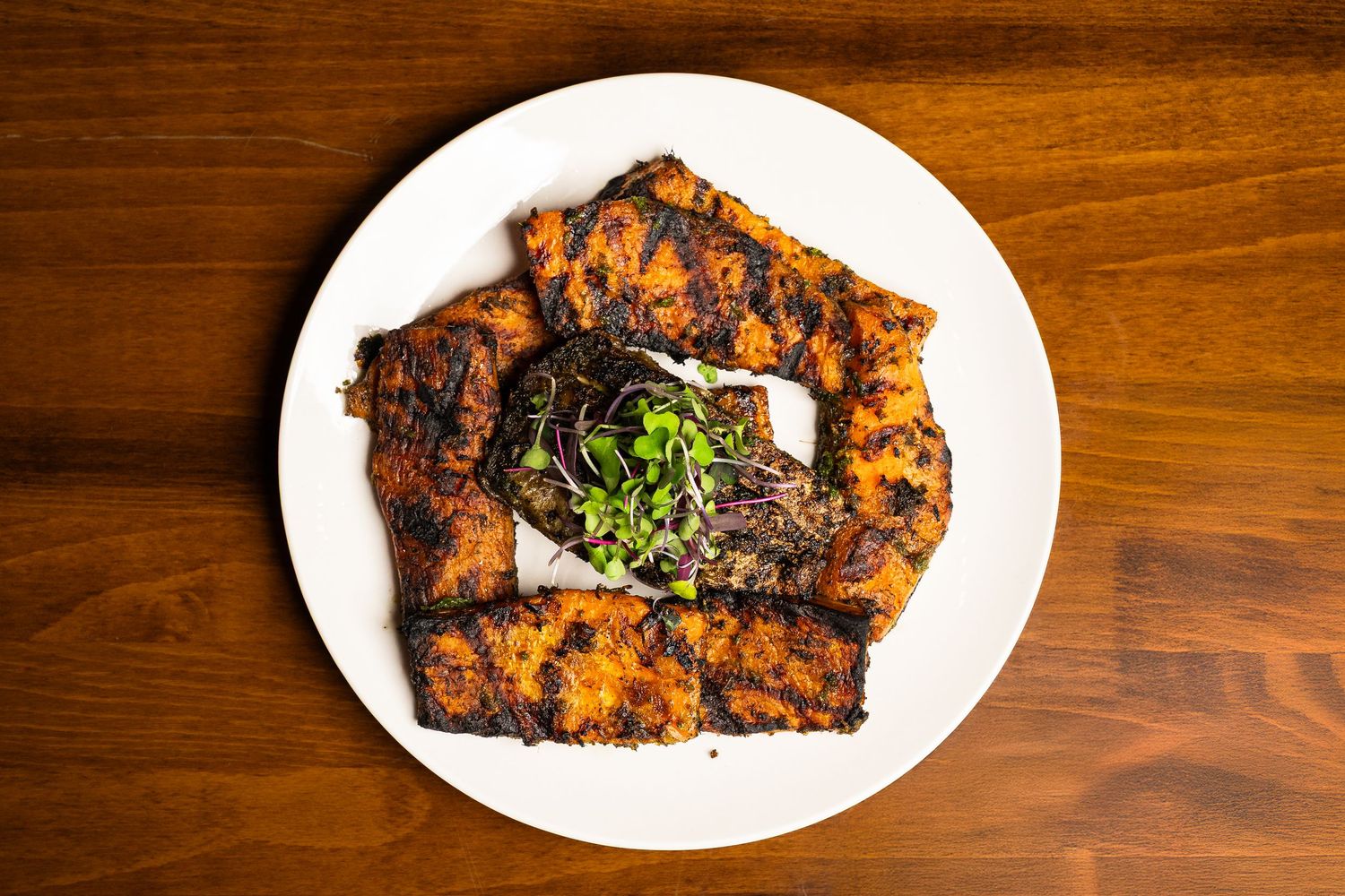 Grilled spiced meat slices arranged on a white plate, garnished with fresh microgreens on a wooden table.