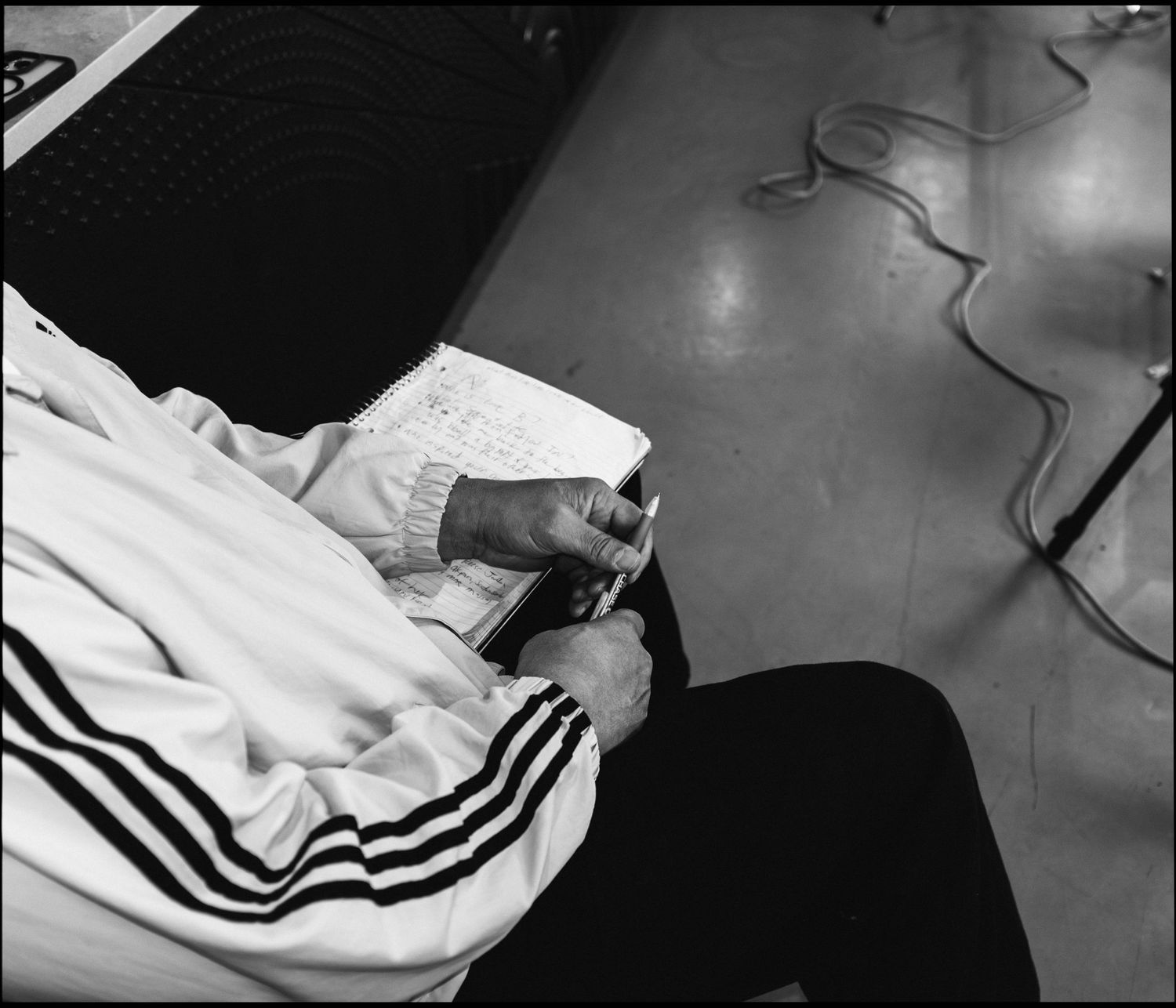 Black and white photo of a person in an Adidas jacket writing in a notebook while seated.
