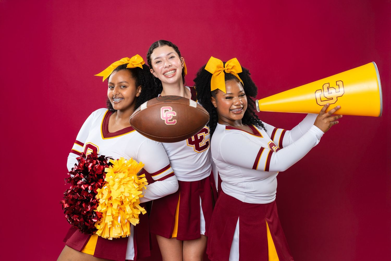 Female soccer team members in maroon uniforms pose with pom poms and megaphones against a red backdrop.