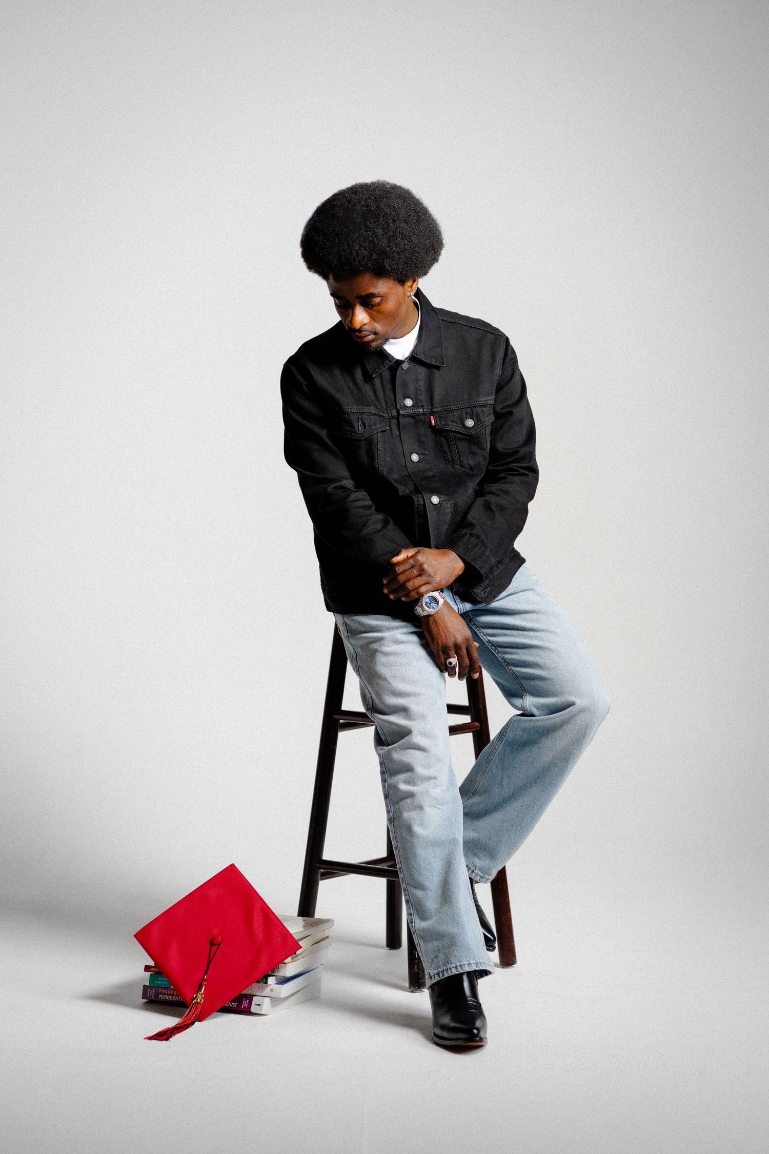 Young Black man with afro in black denim jacket and light jeans sits on stool near red graduation cap.