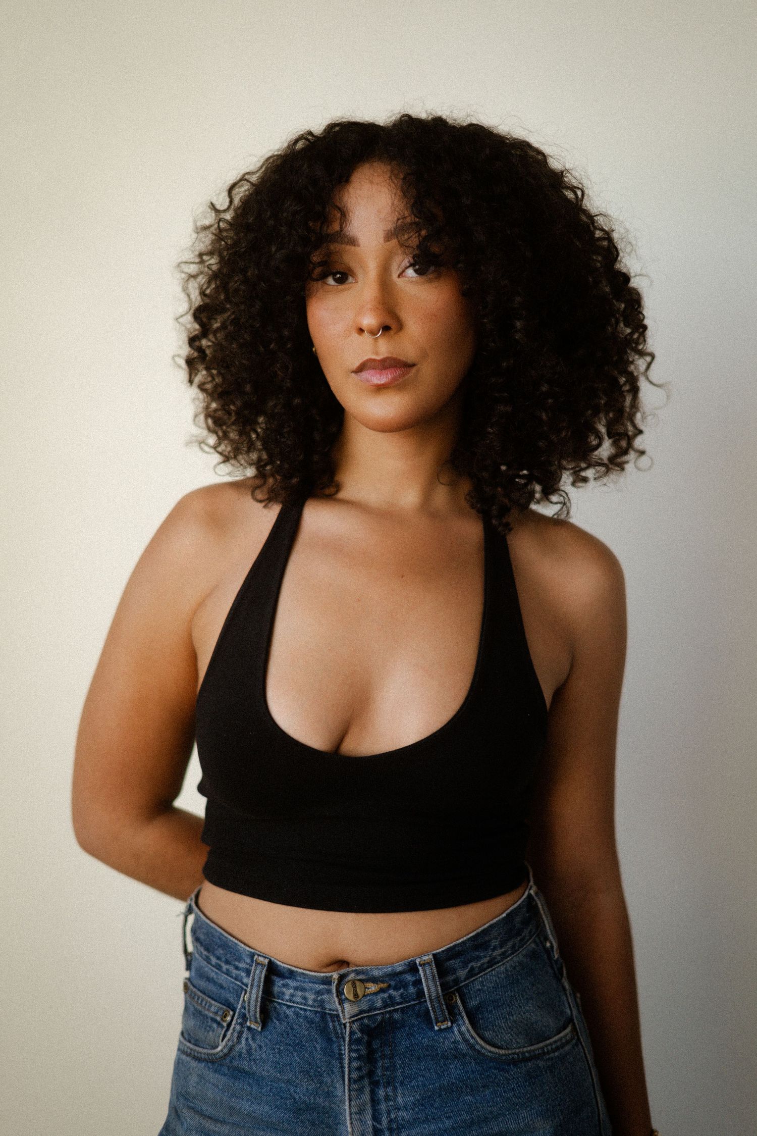 Woman in black tank top and jeans posing confidently against light background.