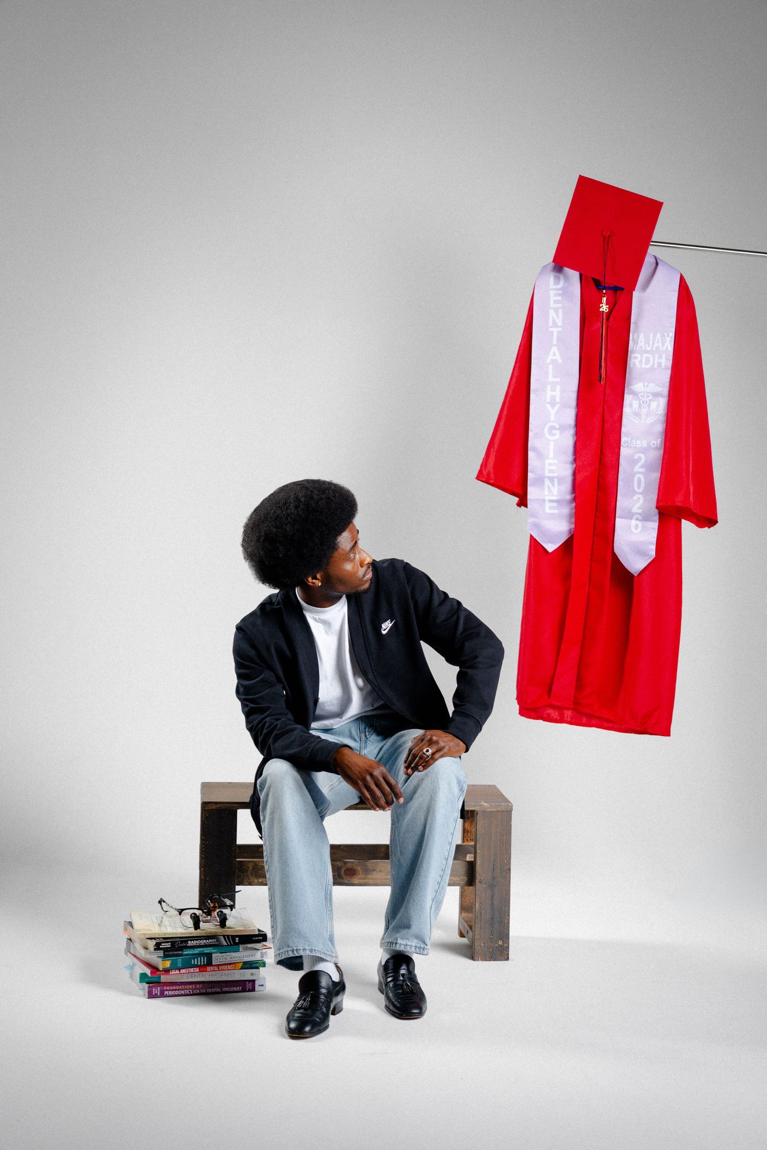 Young man sitting on wooden bench beside stacked books, gazing at red graduation cap and gown hanging nearby.