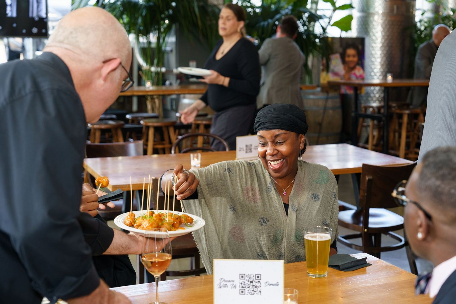 A waiter serves colorful appetizers to a smiling woman at a restaurant table with beer glasses nearby.