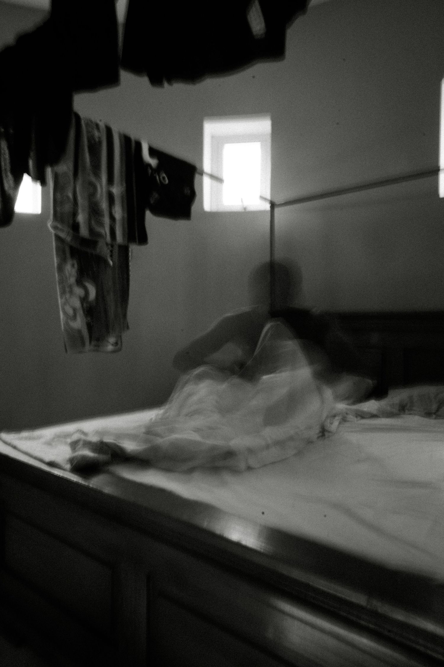 Unmade bed with white sheets in a dimly lit room with clothes hanging above.