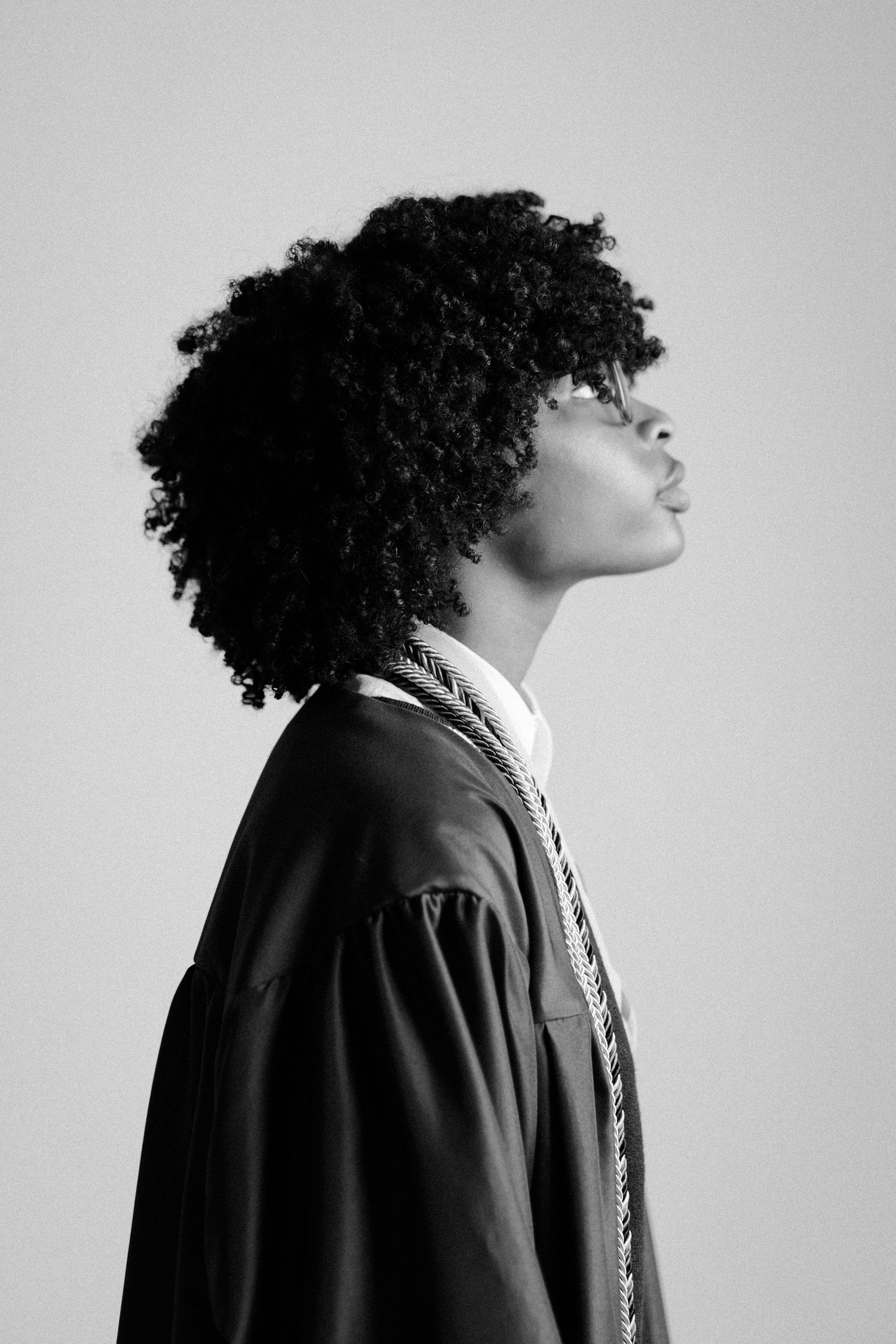 Black and white profile portrait of a young person with curly hair wearing a graduation gown and honor cords.