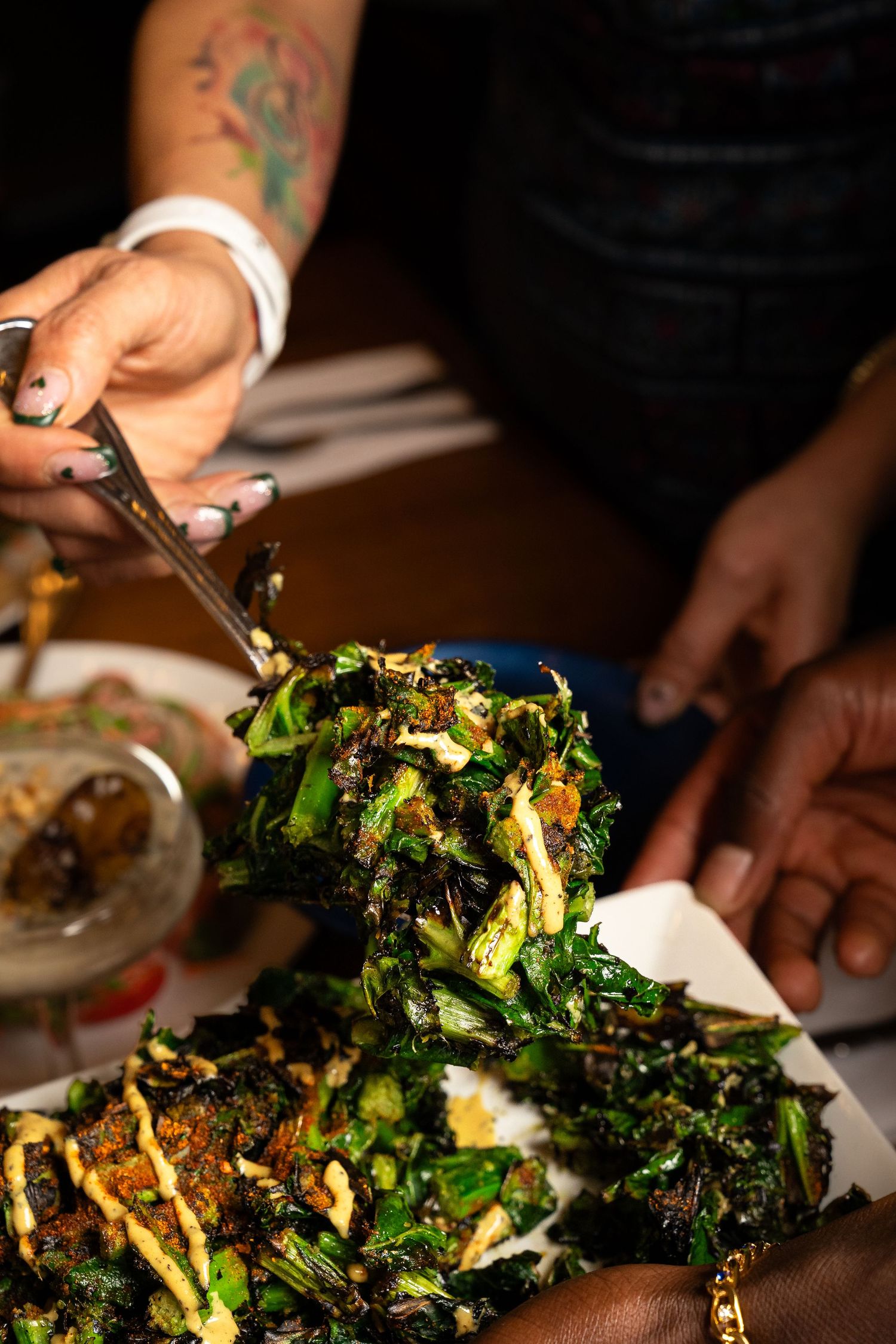 Hands serving crispy sautéed greens with mustard drizzle using tongs at dinner.