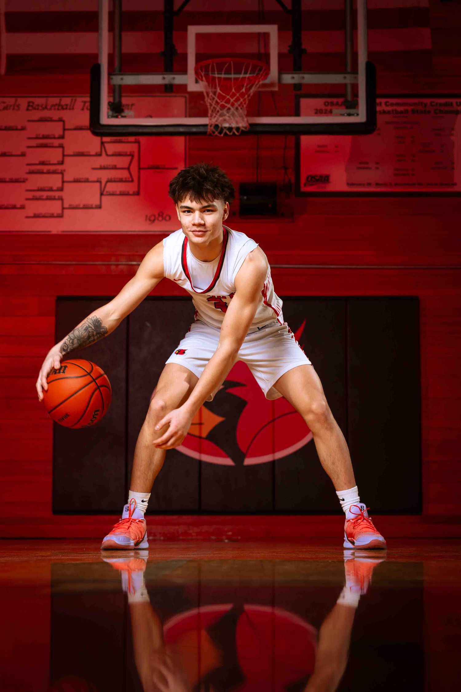 Basketball player crouches in defensive stance on red-lit court wearing white jersey and coral shoes.