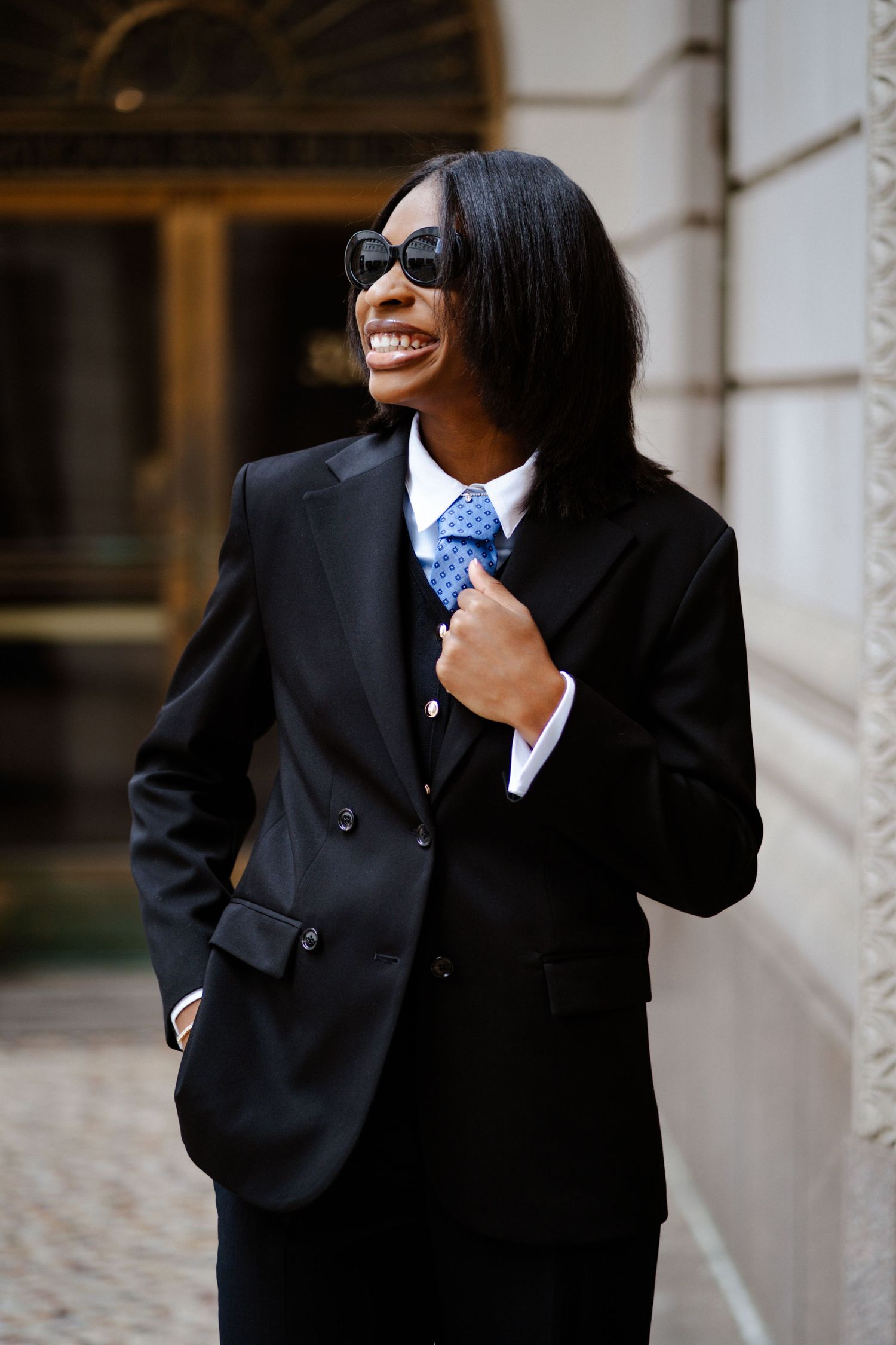 Smiling Black businesswoman in black suit and blue tie wearing sunglasses outdoors near building.