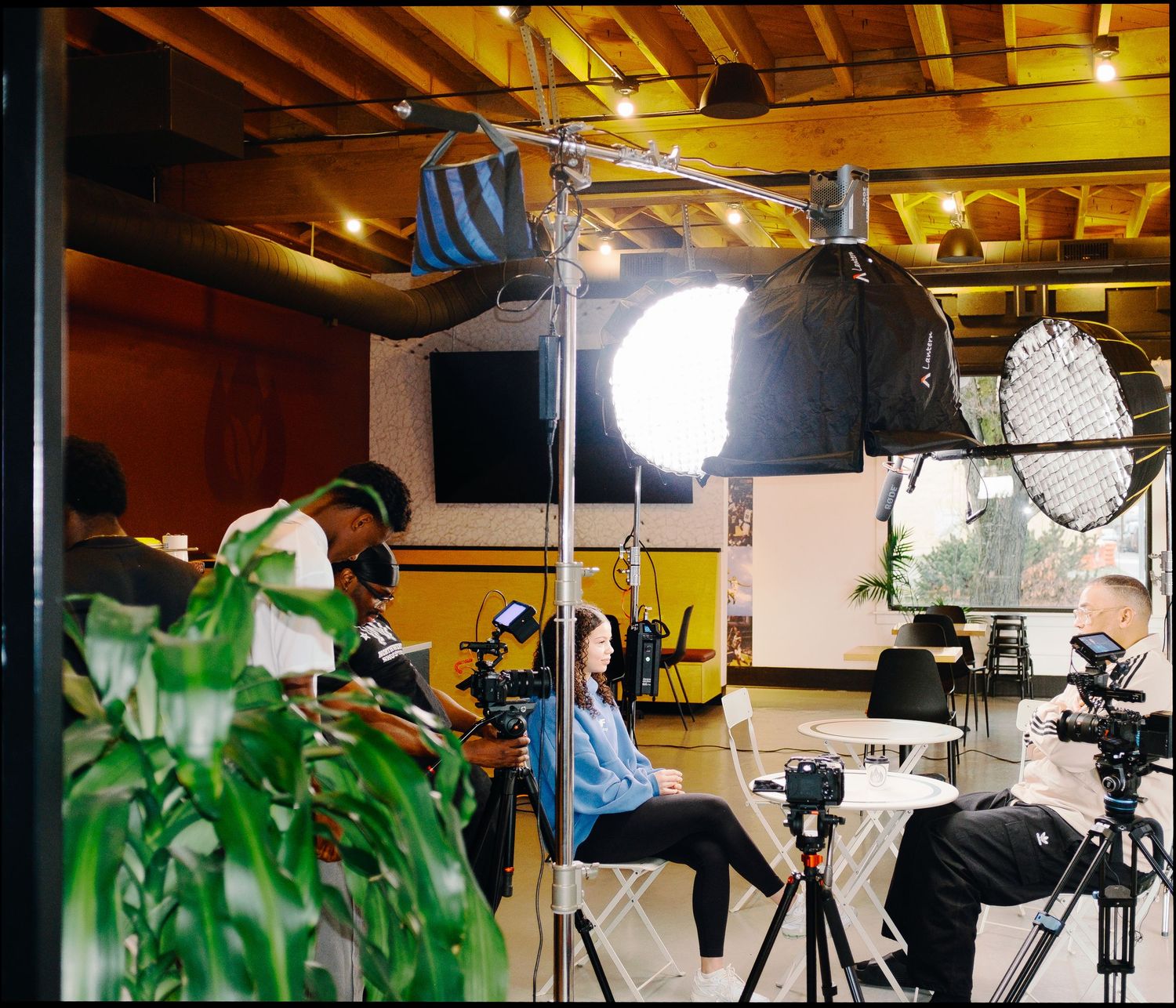 Film crew sets up professional studio lighting equipment for an indoor video production shoot with seated subject.