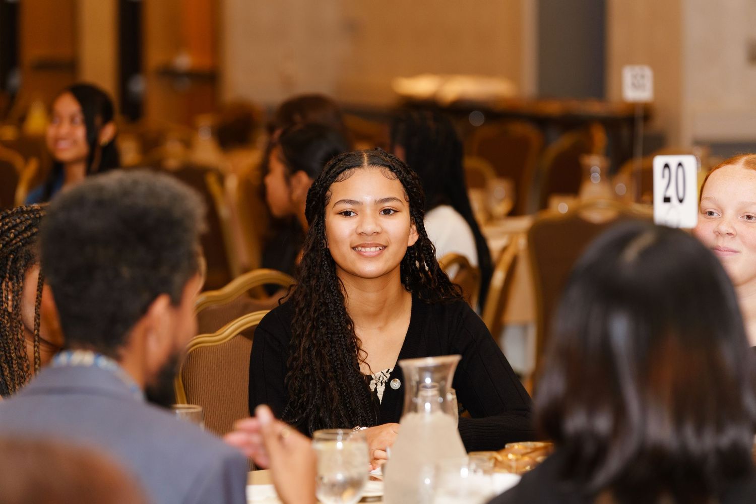 Young woman with braided hair smiles at a formal banquet dinner event, seated at a table numbered 20.