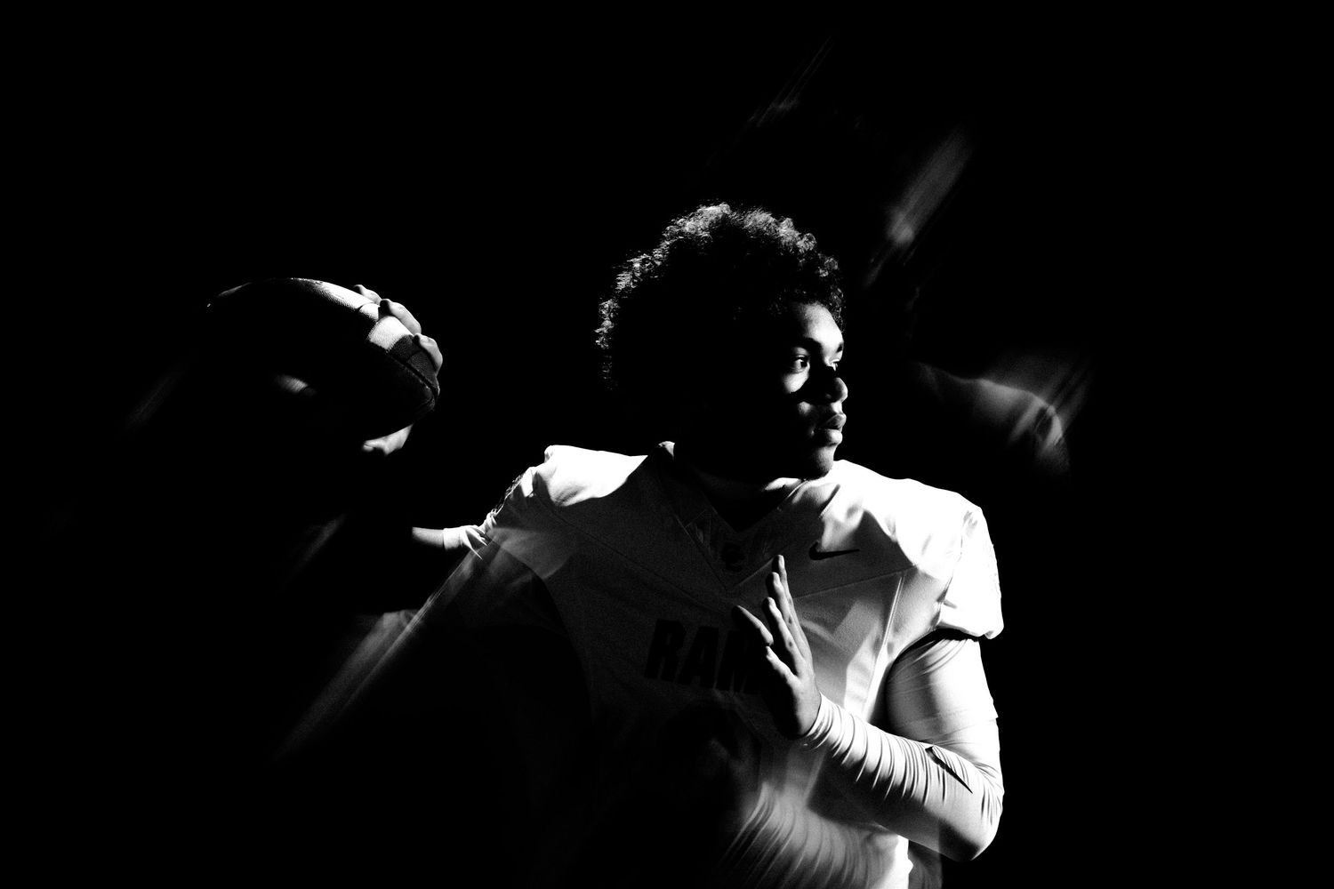 Black and white photo of a football player in white uniform holding a ball in dramatic low-key lighting.