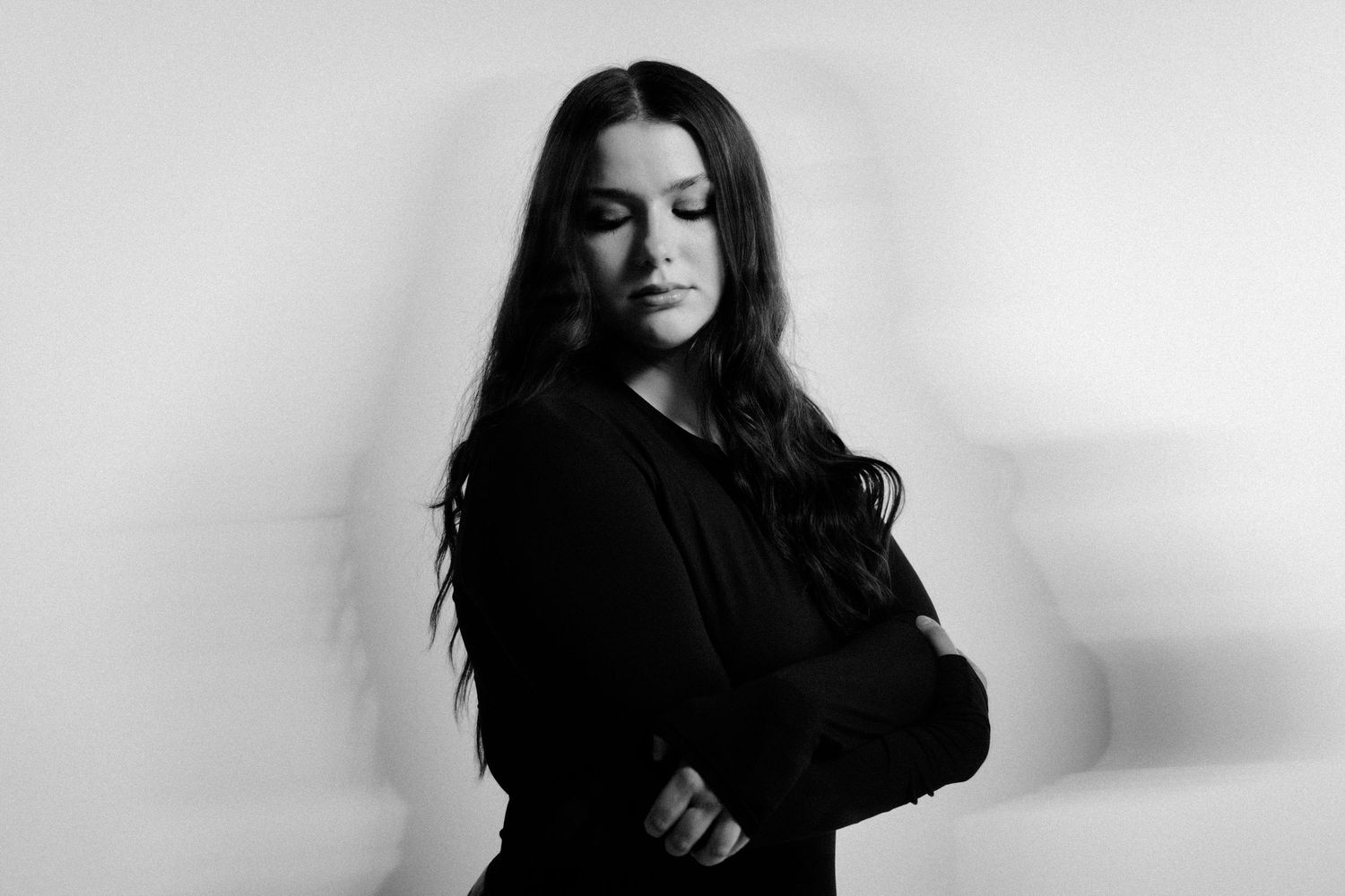 Black and white portrait of a woman with long dark hair in a black outfit, arms crossed, looking down thoughtfully.