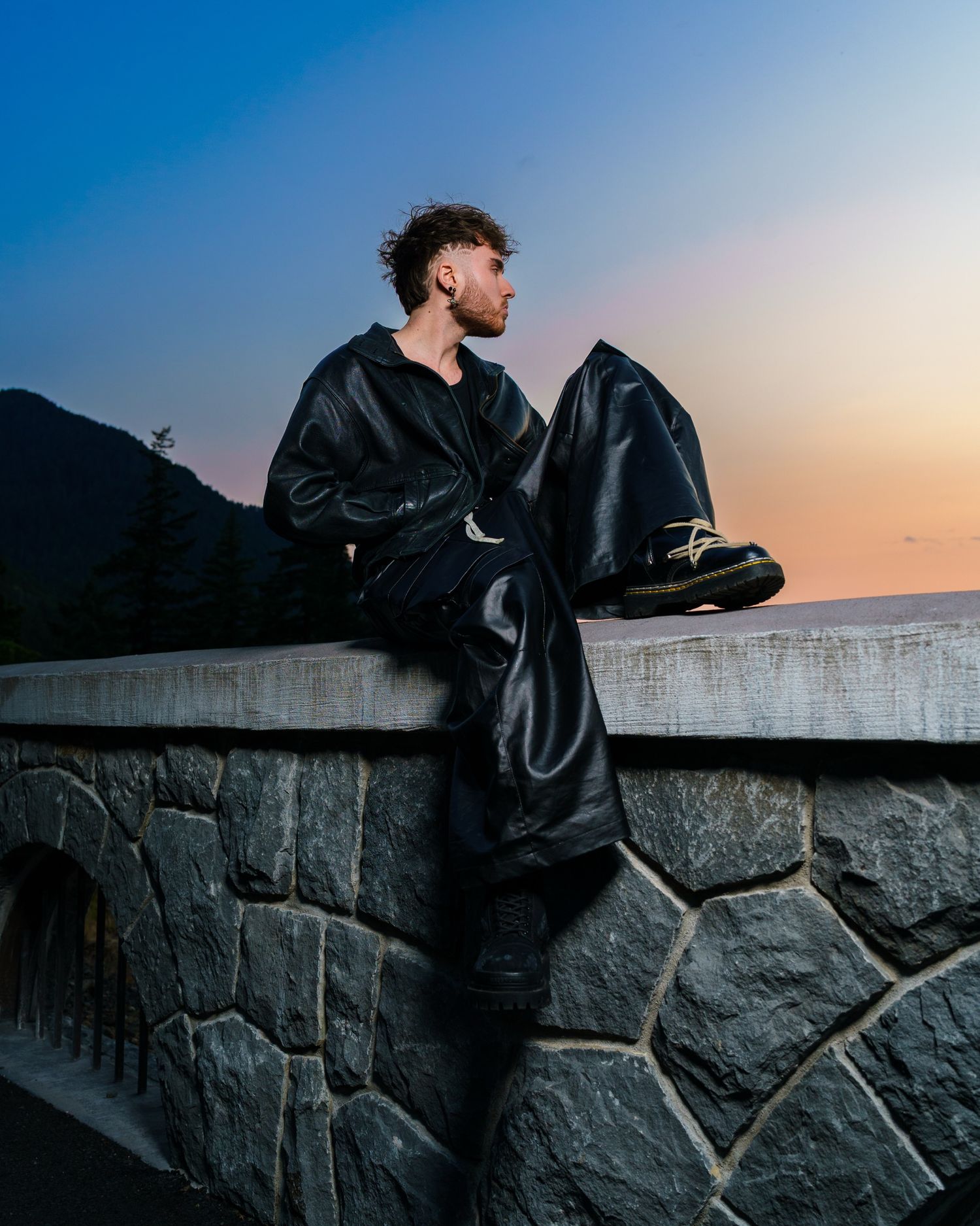 Figure in leather jacket leaning against stone wall during sunset.