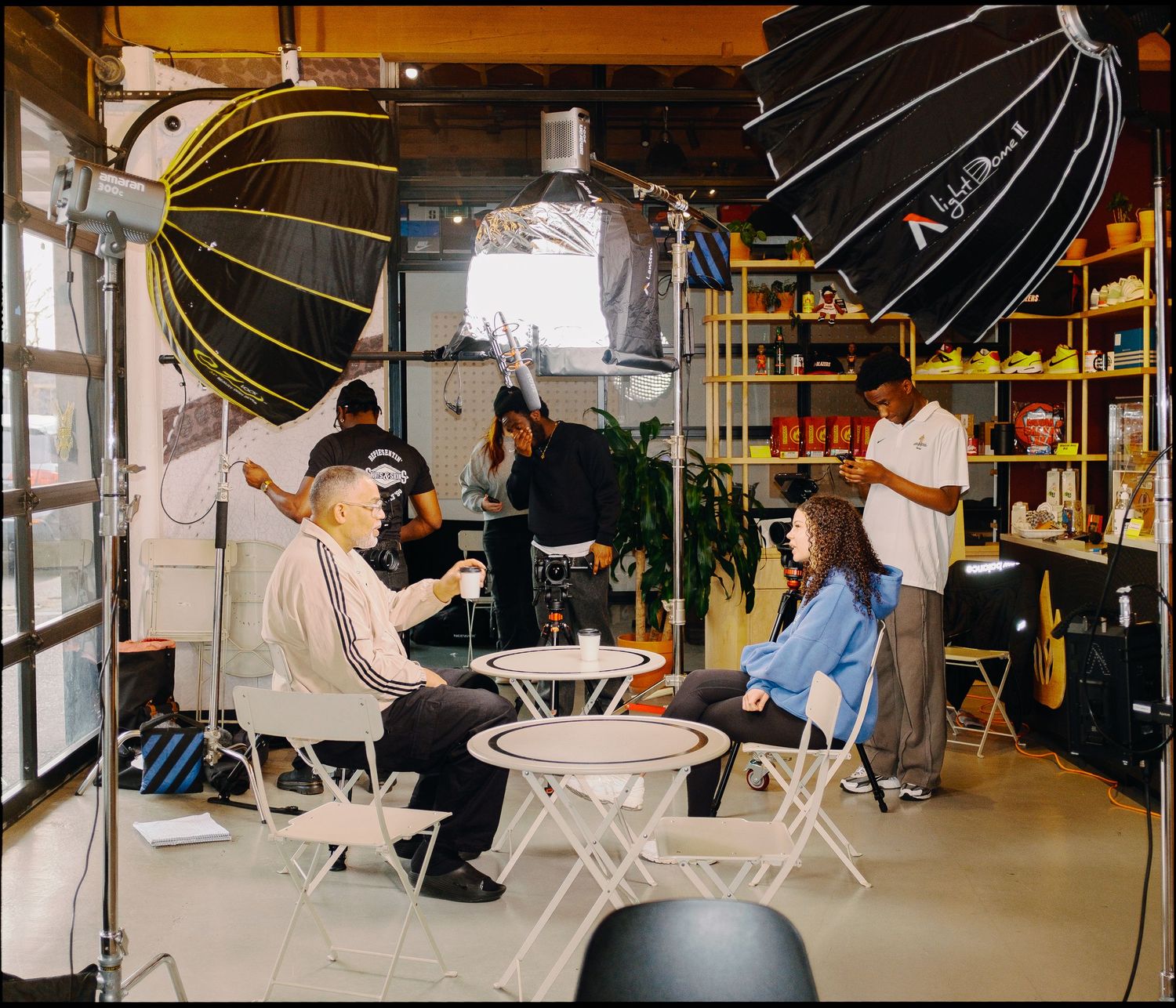 Photography studio setup with softbox lights, people sitting at white folding tables, and shelves of supplies in background.