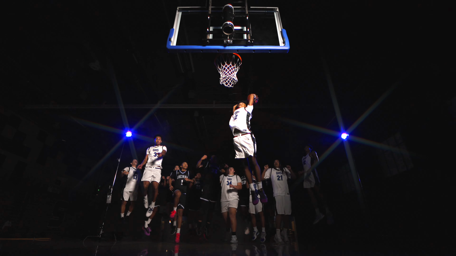 Basketball player dunks under dramatic blue lights in a dark arena with teammates watching from below.