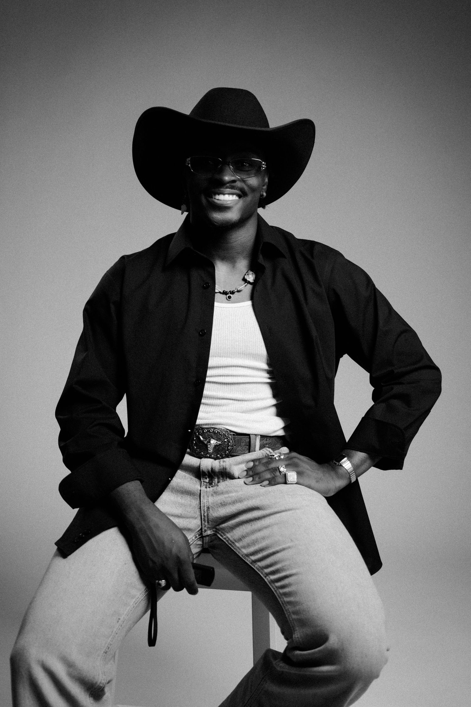 Smiling man in black cowboy hat, sunglasses, black shirt, white tank, and jeans poses seated in black and white photo.