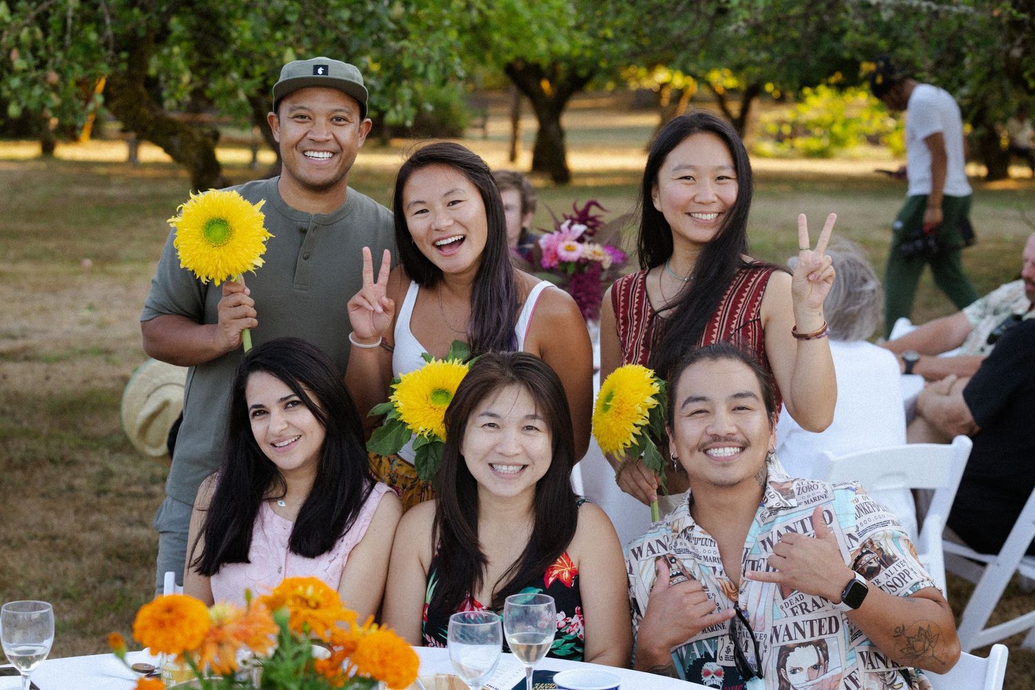 Group of seven friends smiling and posing with peace signs at an outdoor garden dinner party with sunflowers.