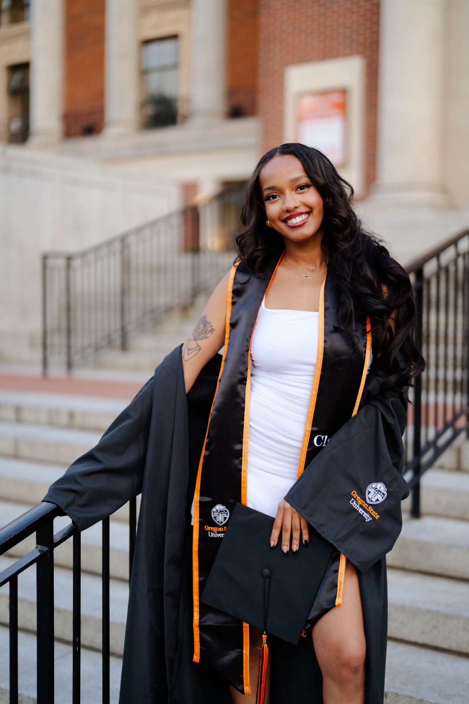 Smiling graduate in black cap and gown with orange stole poses on university steps in white dress.