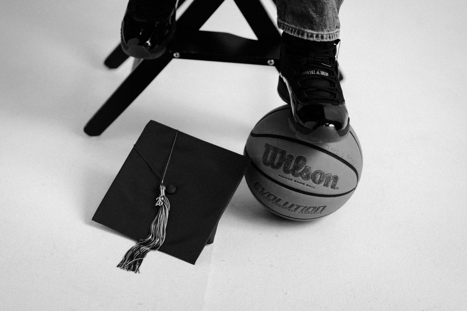 Black and white photo of a Wilson basketball with a graduation cap beside it and a sneaker resting on top.