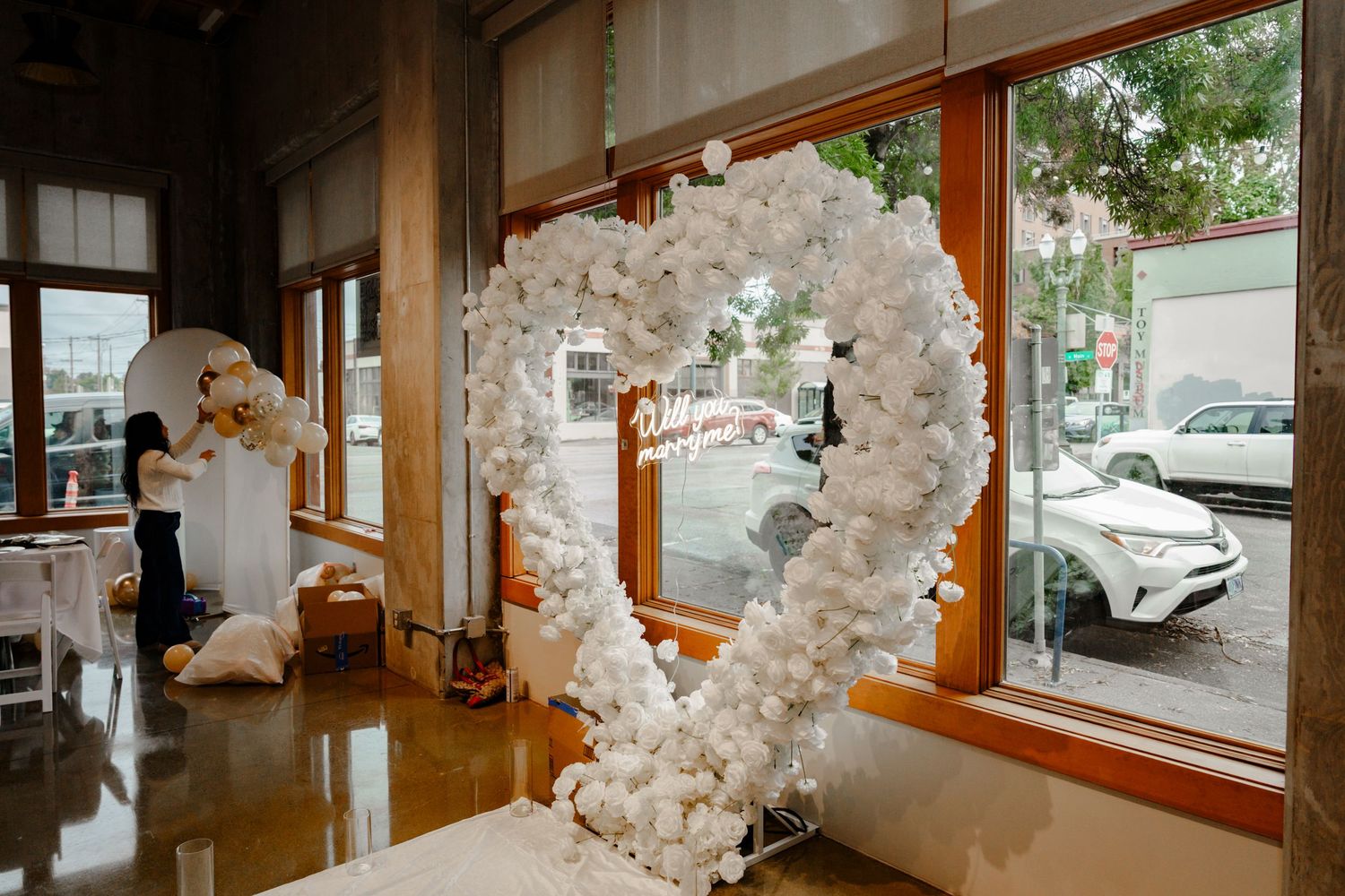 White floral heart arch with mirror sign displayed in elegant wedding venue with large windows.