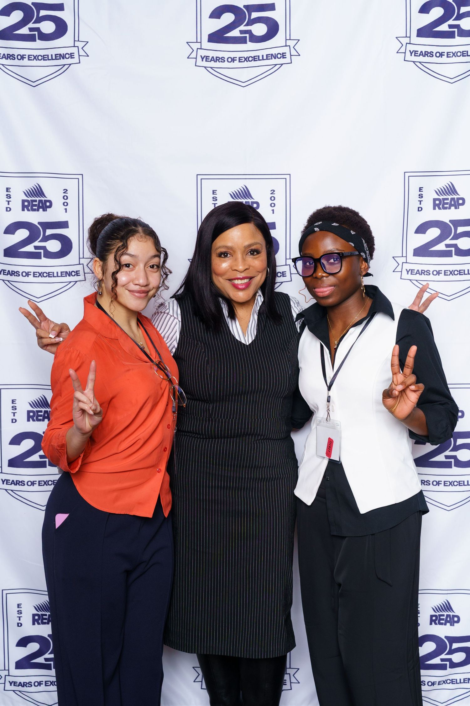 Three women pose with peace signs at REAP 25 Years of Excellence celebration event backdrop.