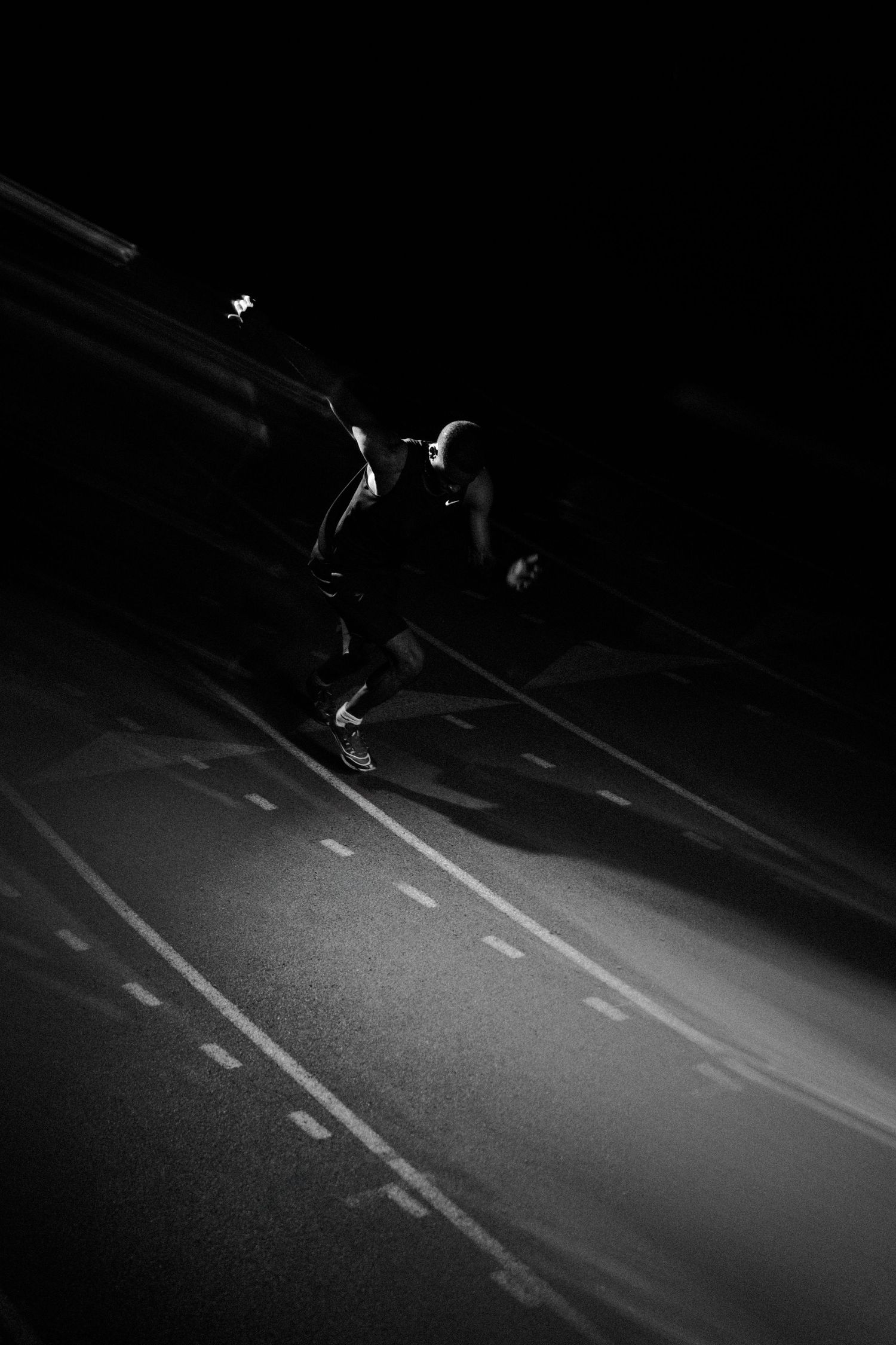 Black and white photo of a runner sprinting on a track at night, illuminated by a single light source.