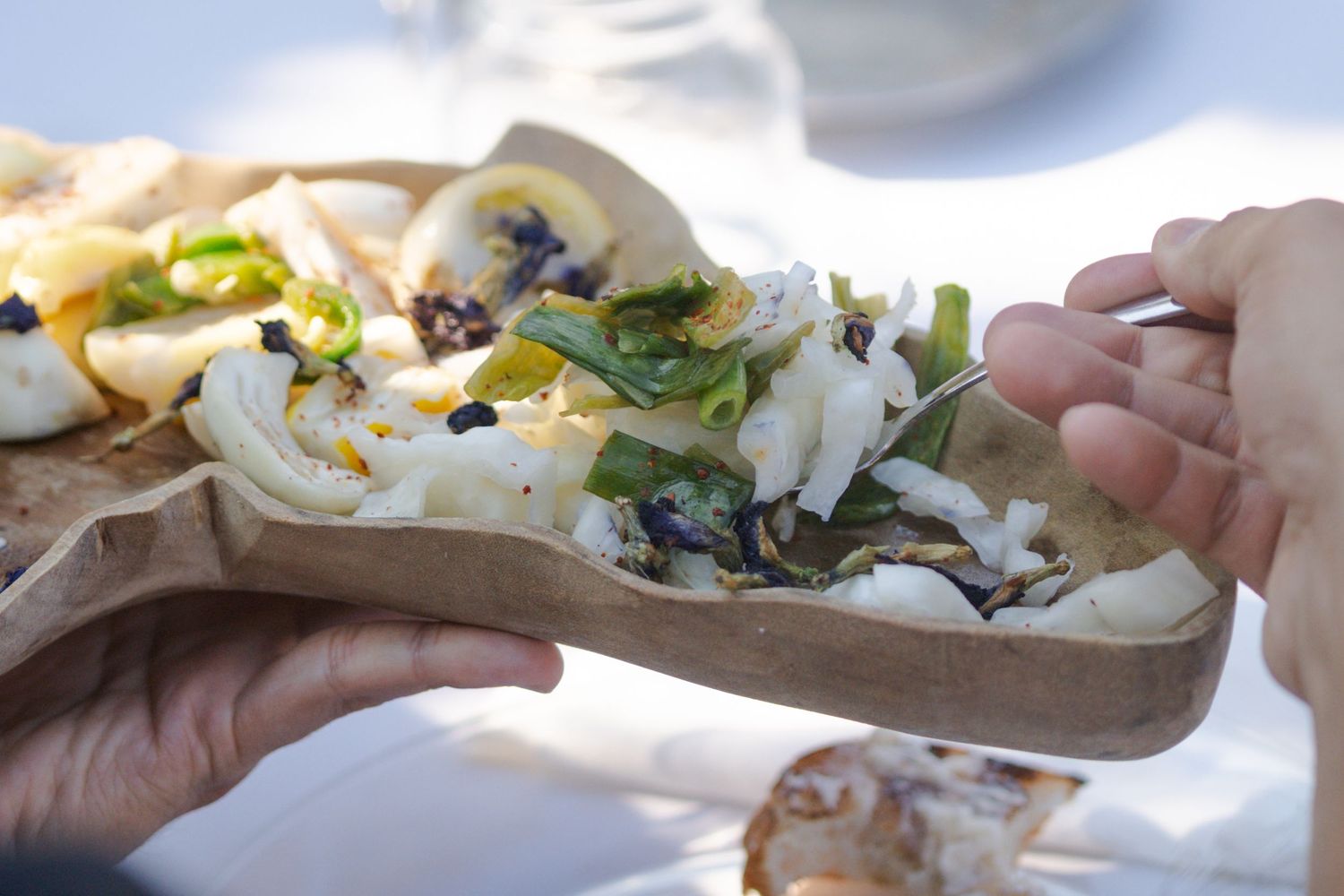 Hands holding a wooden platter with fresh salad topped with greens, edible flowers, and white cheese outdoors.