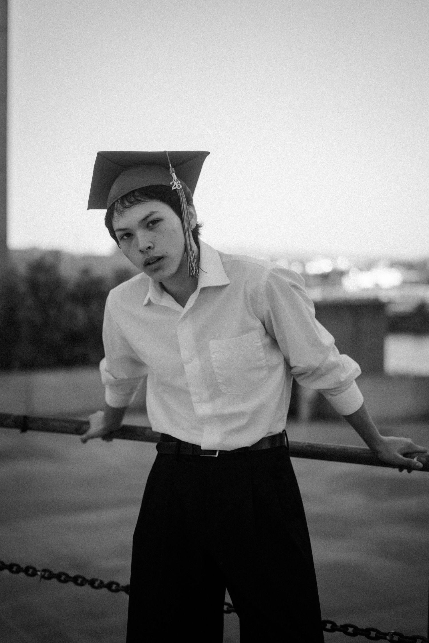 Black and white photo of a young person in graduation cap and white shirt leaning on a chain railing outdoors.