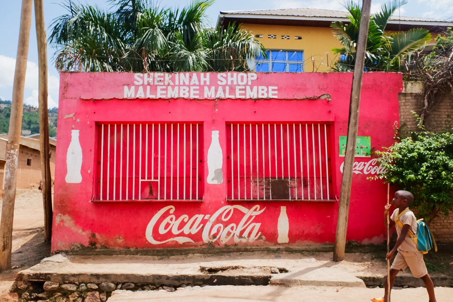 Long red Coca-Cola branded wall with palm trees in an African village setting.