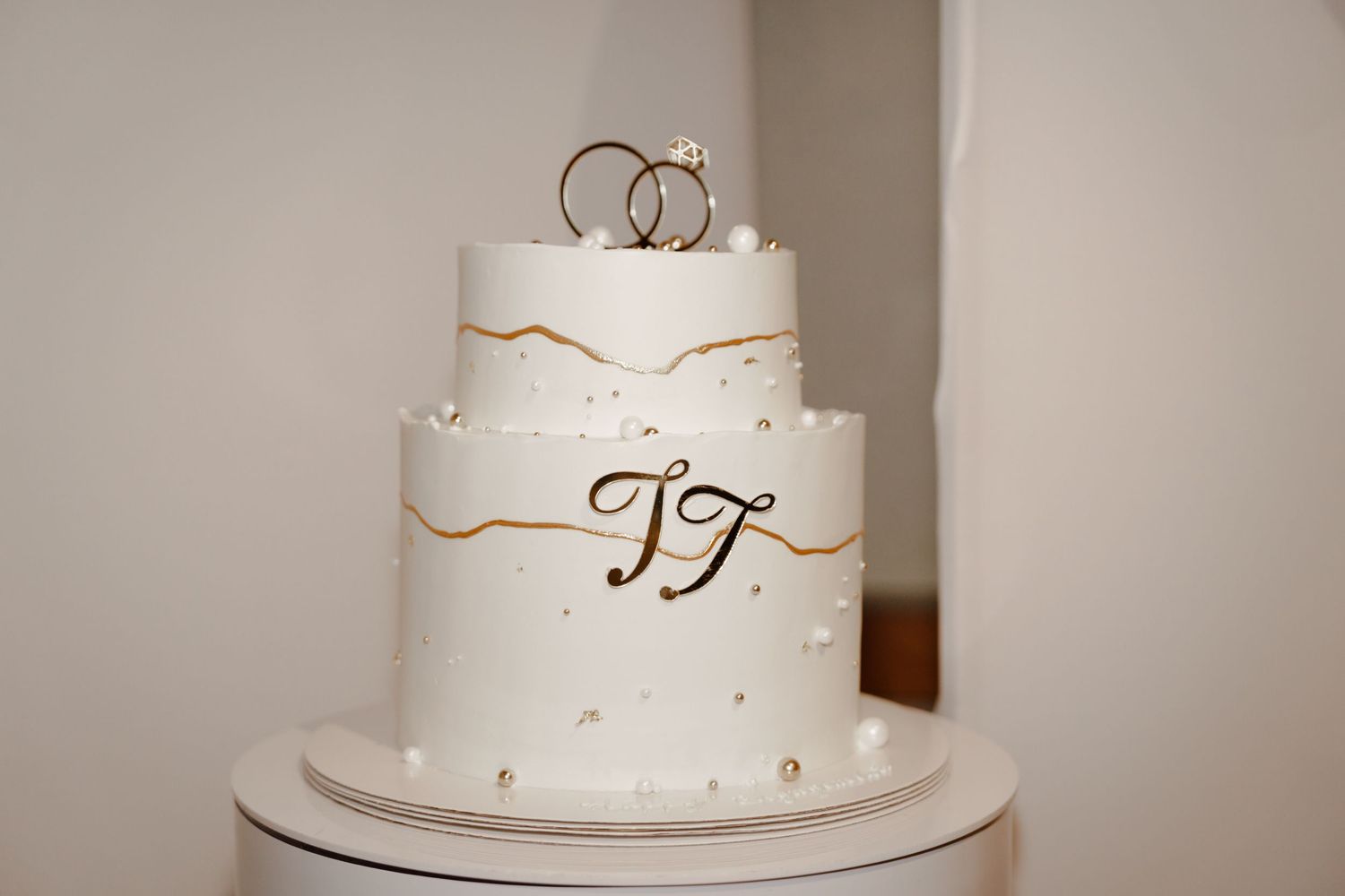 Elegant two-tier white wedding cake with gold monogram TT, pearl accents, and wedding rings topper.