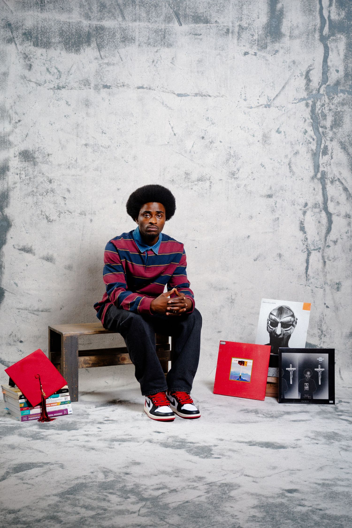 Young man with afro sits on bench surrounded by vinyl records and books against gray concrete wall.