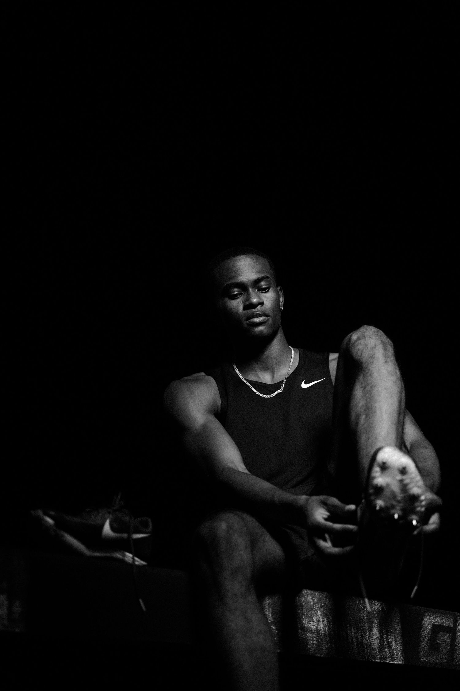 Black and white photo of a muscular athlete in Nike tank top sitting in dramatic low-key lighting.