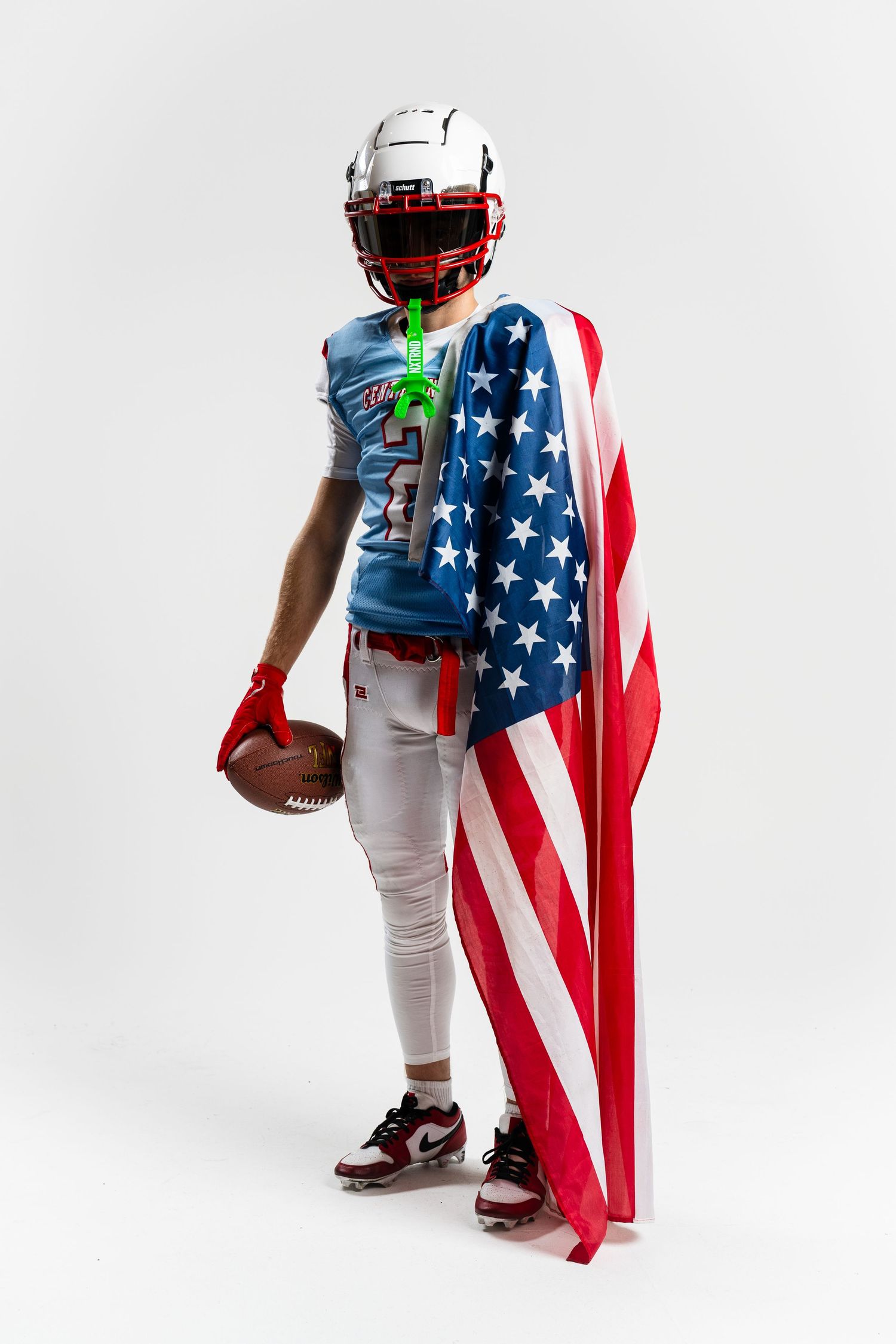 Football player poses with American flag draped as cape against white background.