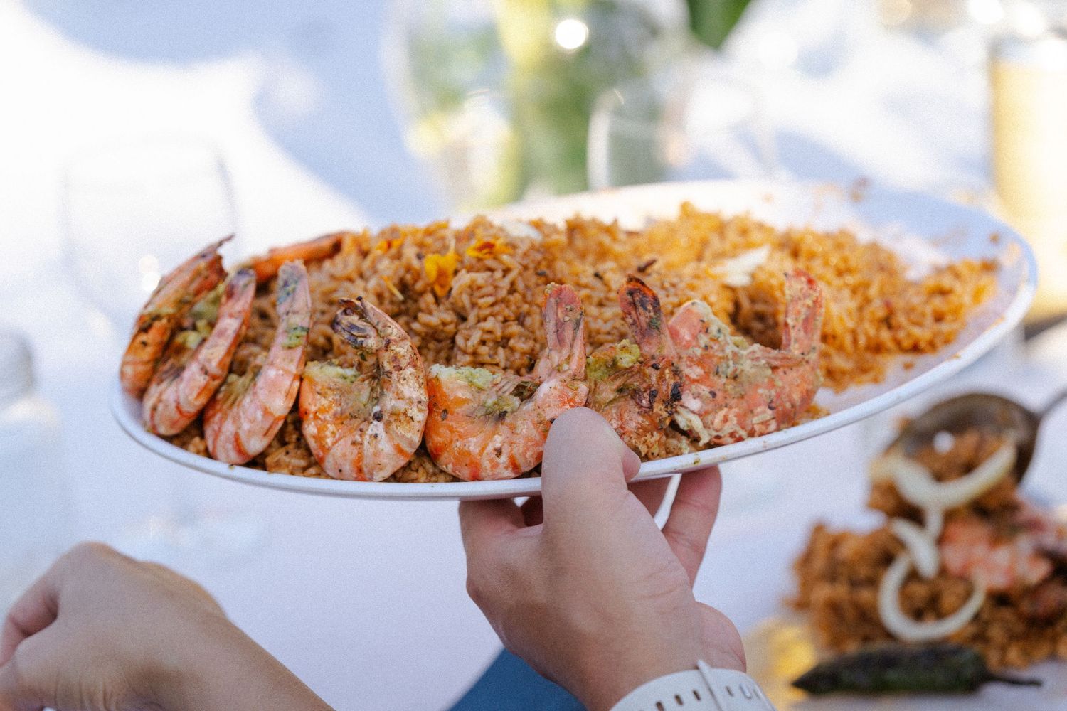 Hands holding a large oval platter of shrimp over seasoned rice at an elegant outdoor dining event.