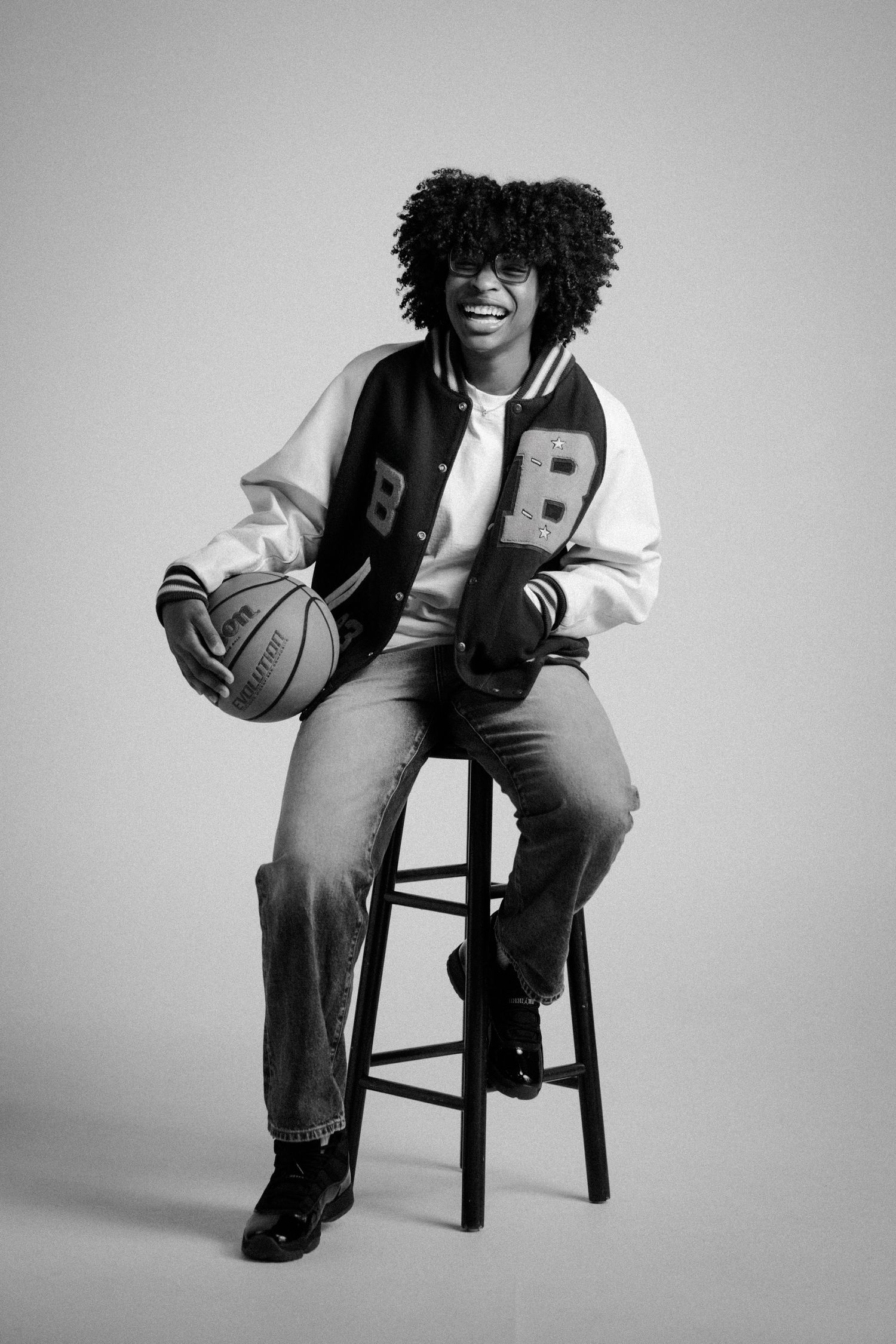 Smiling person in varsity jacket holding basketball, seated on stool, black and white portrait photo.