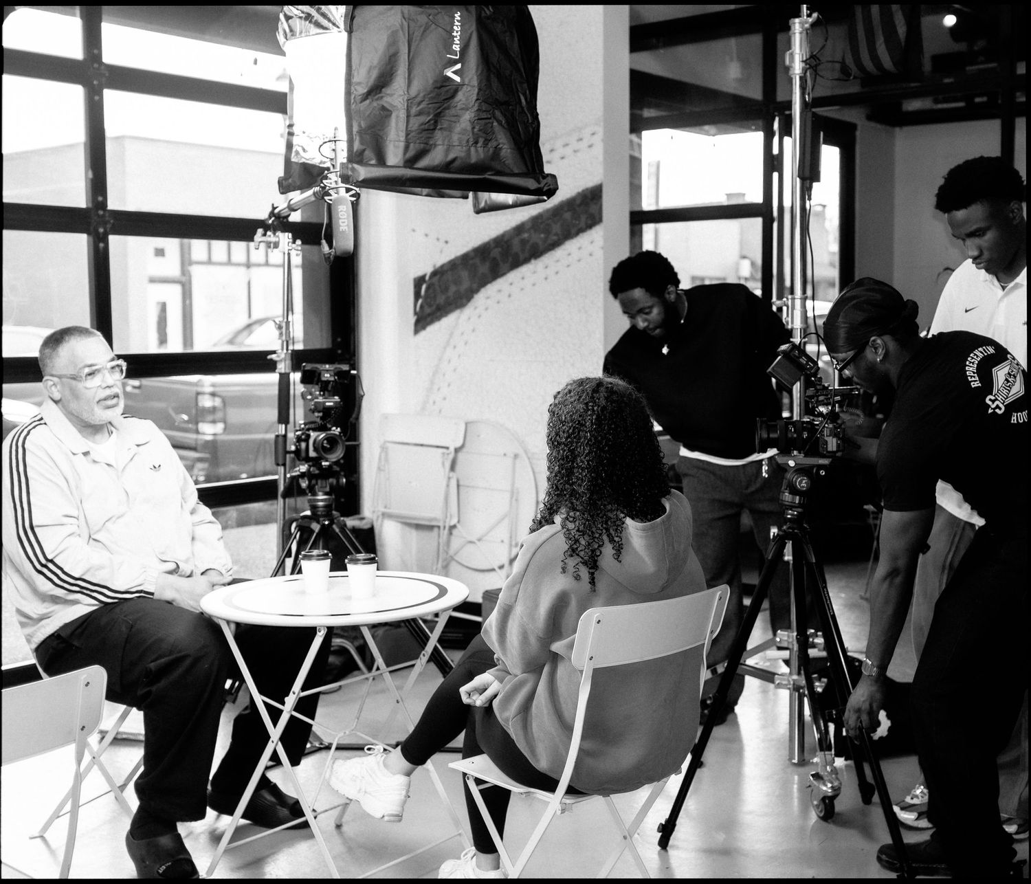 Black and white photo of a film crew interviewing a man at a small table with cameras and lighting equipment.
